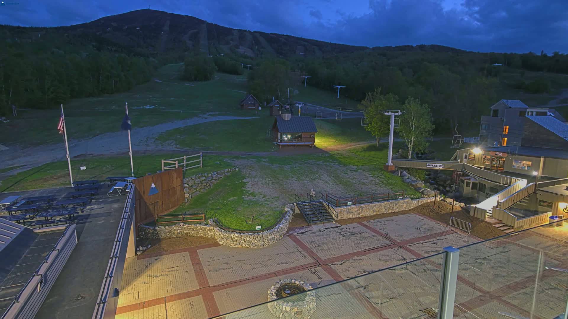 A nighttime view of a ski resort with illuminated buildings and a paved area, under a partly cloudy sky, with a mountain in the background.