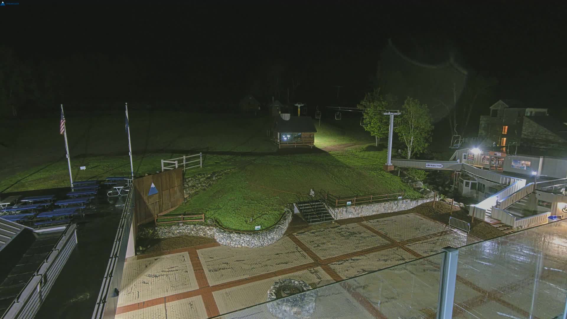 A nighttime view of an outdoor plaza area with a stone retaining wall, a small building, a ski lift, and picnic tables, illuminated by artificial lights.