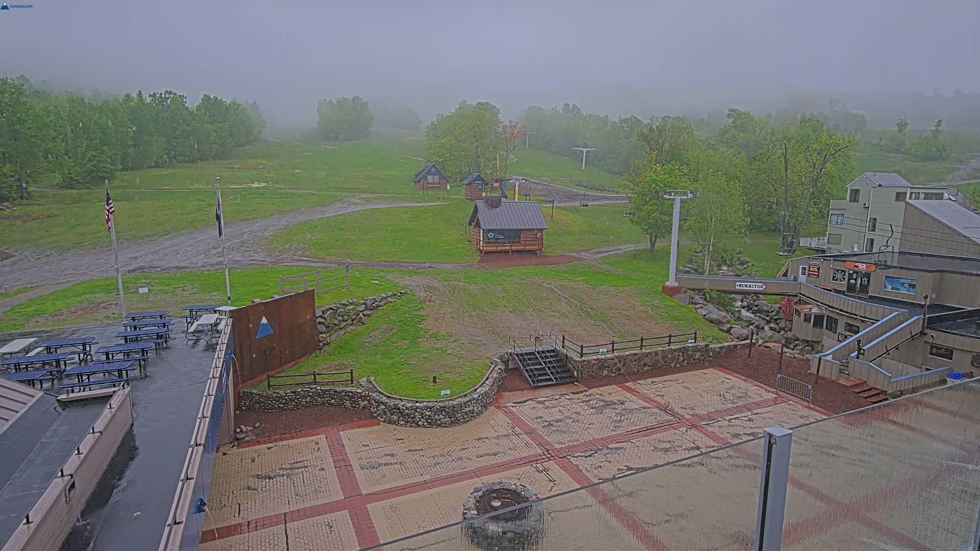 A rain-soaked ski resort base area with several small buildings, picnic tables, and a paved plaza, surrounded by green hills and trees under an overcast sky.