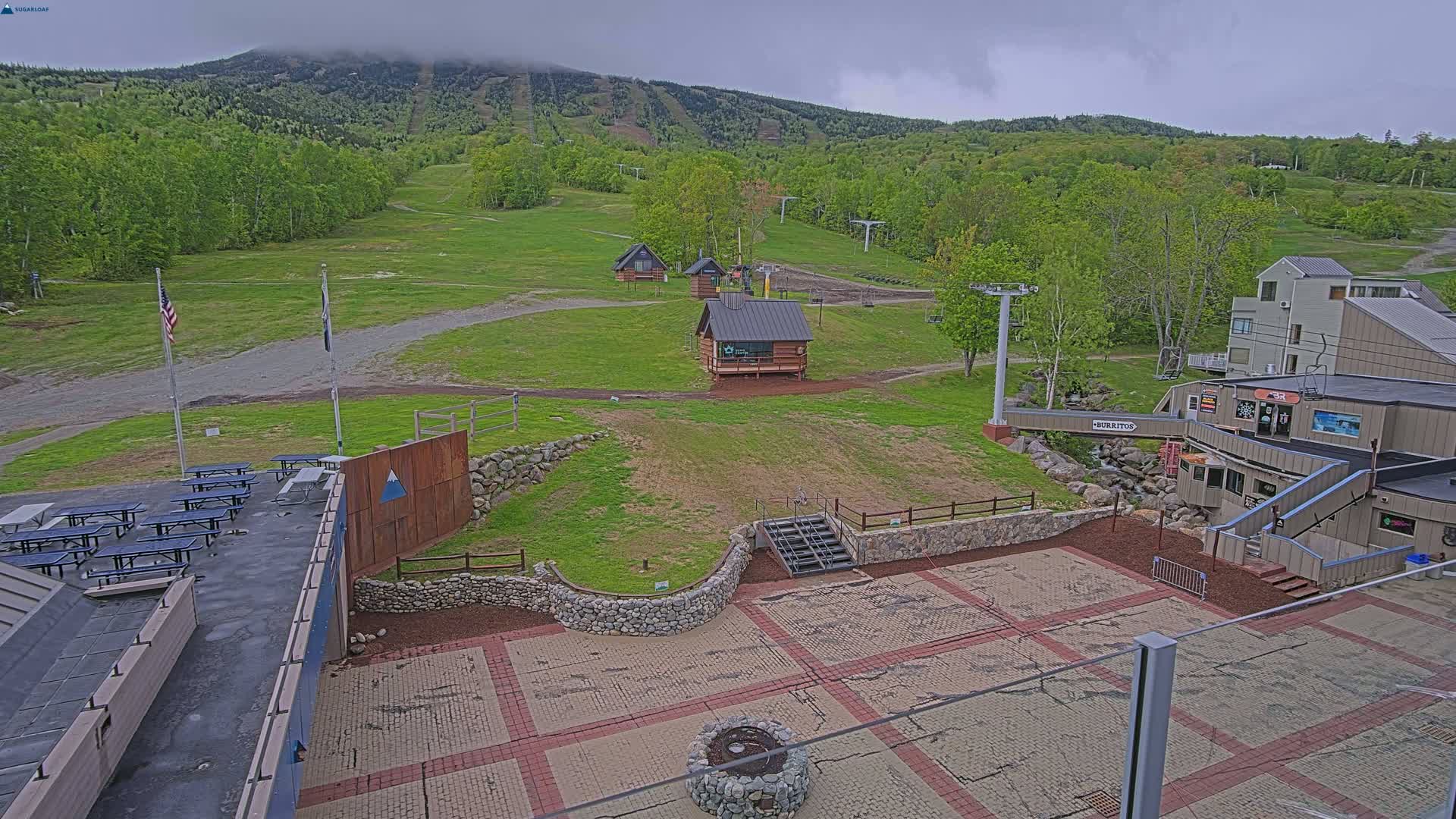 A paved plaza with picnic tables and a stone fire pit overlooks grassy ski slopes and a mountain under an overcast sky.