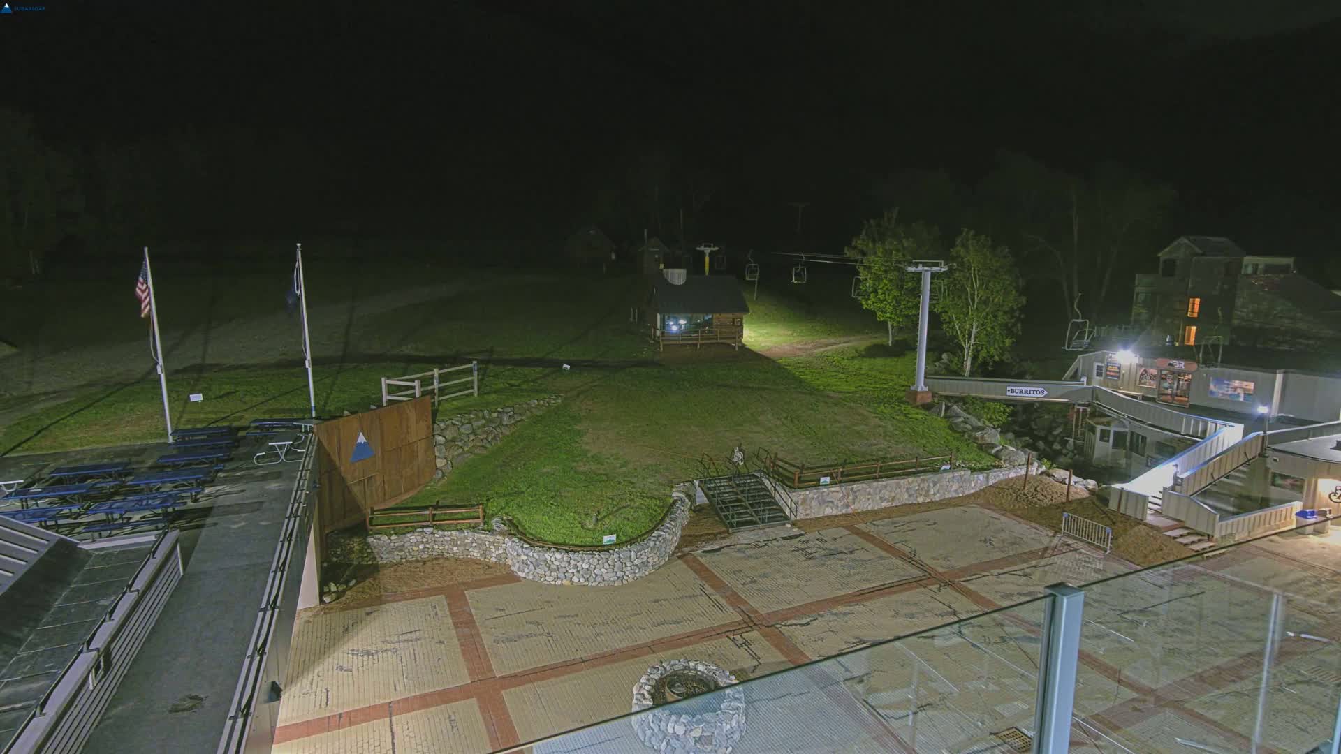 At night, a ski resort's base area is illuminated, showing a grassy area with a stone wall, picnic tables, a small building, and a chairlift.