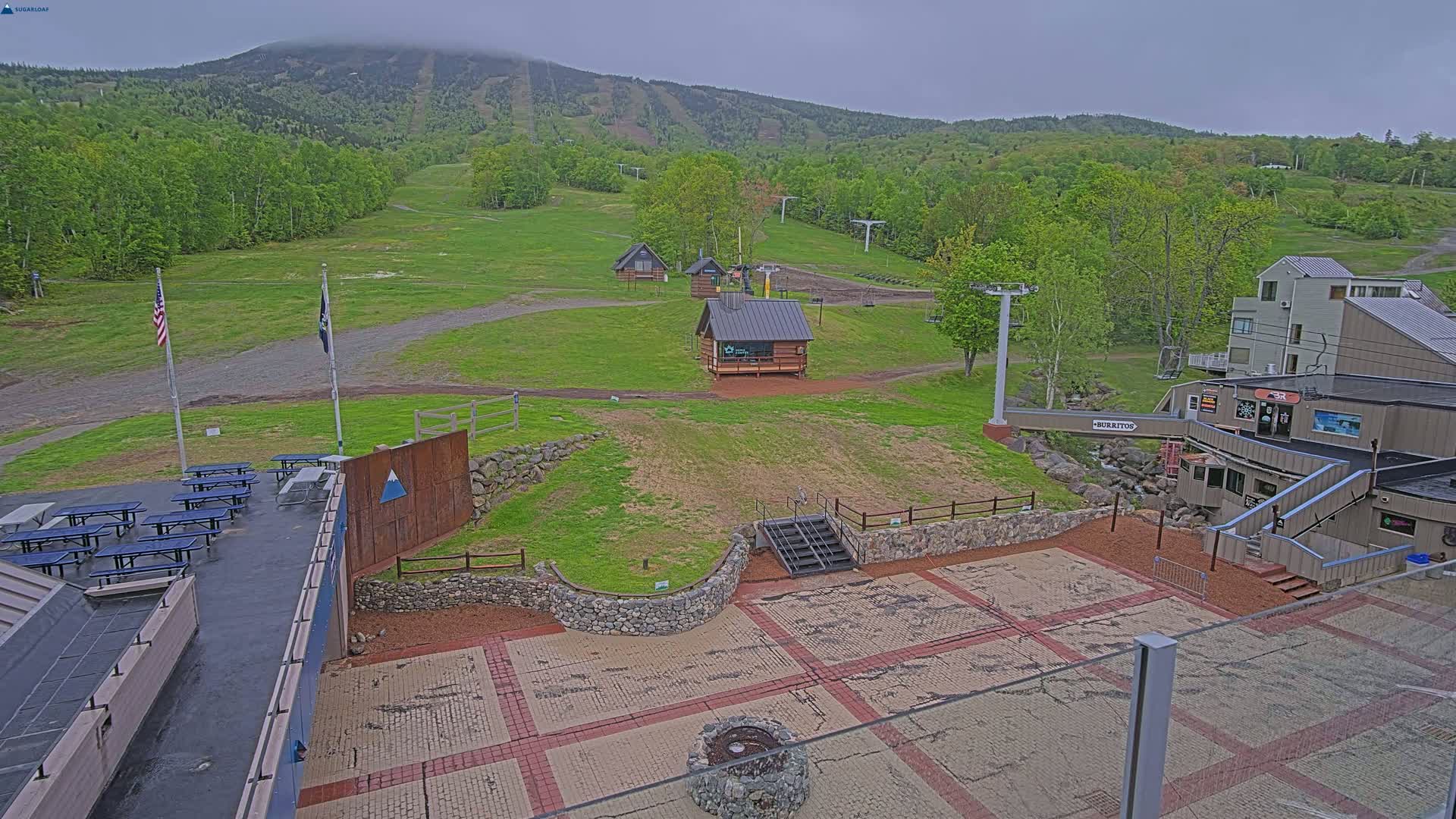 A paved patio area with picnic tables and small buildings overlooks a grassy ski slope and a mountain shrouded in fog.