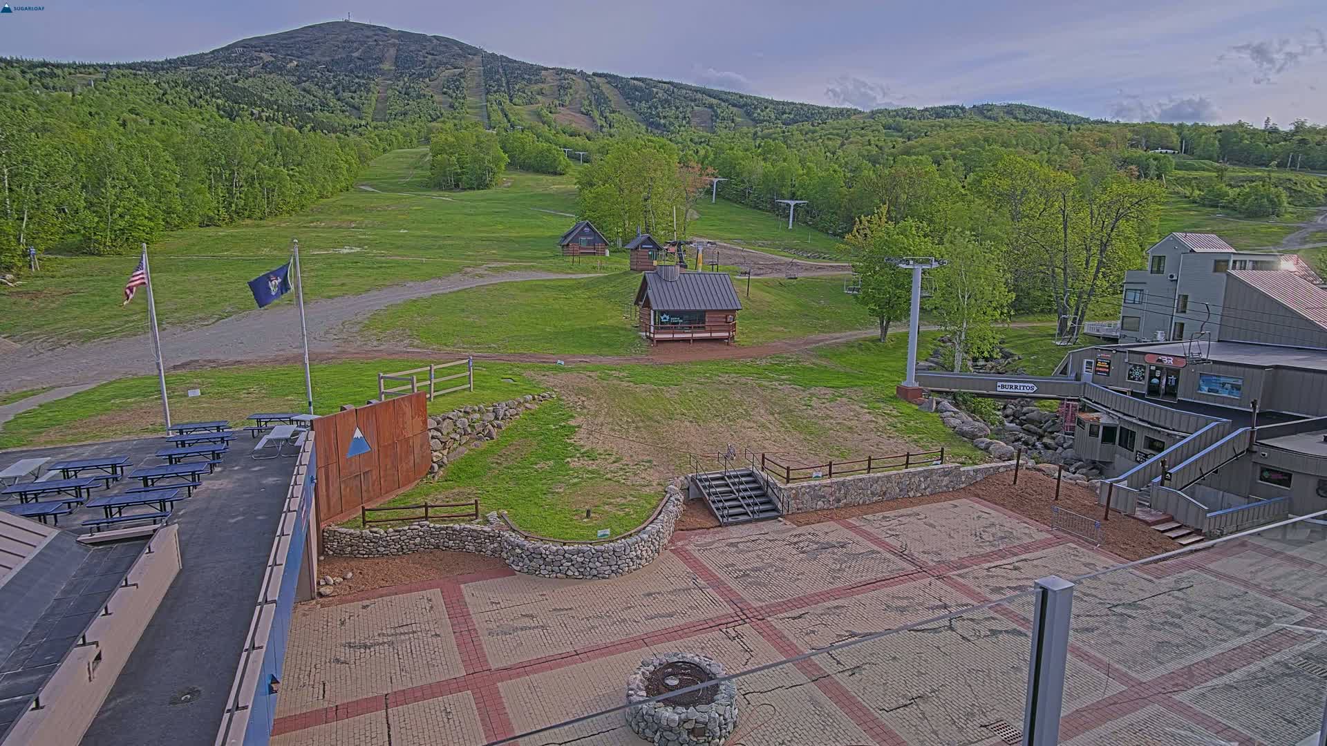 A mostly sunny view of a ski resort in spring, showing grassy slopes, several small buildings, picnic tables, and a paved area with a fire pit, all set against a backdrop of a mountain.