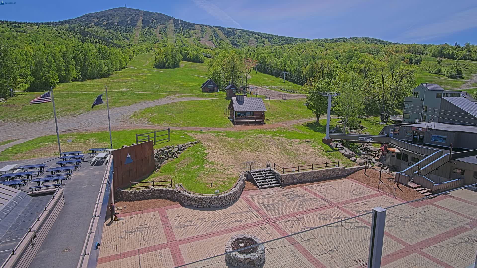 An outdoor patio area with picnic tables, small buildings, and a ski lift overlooks a grassy hillside dotted with trees and ski runs under a bright, sunny sky.