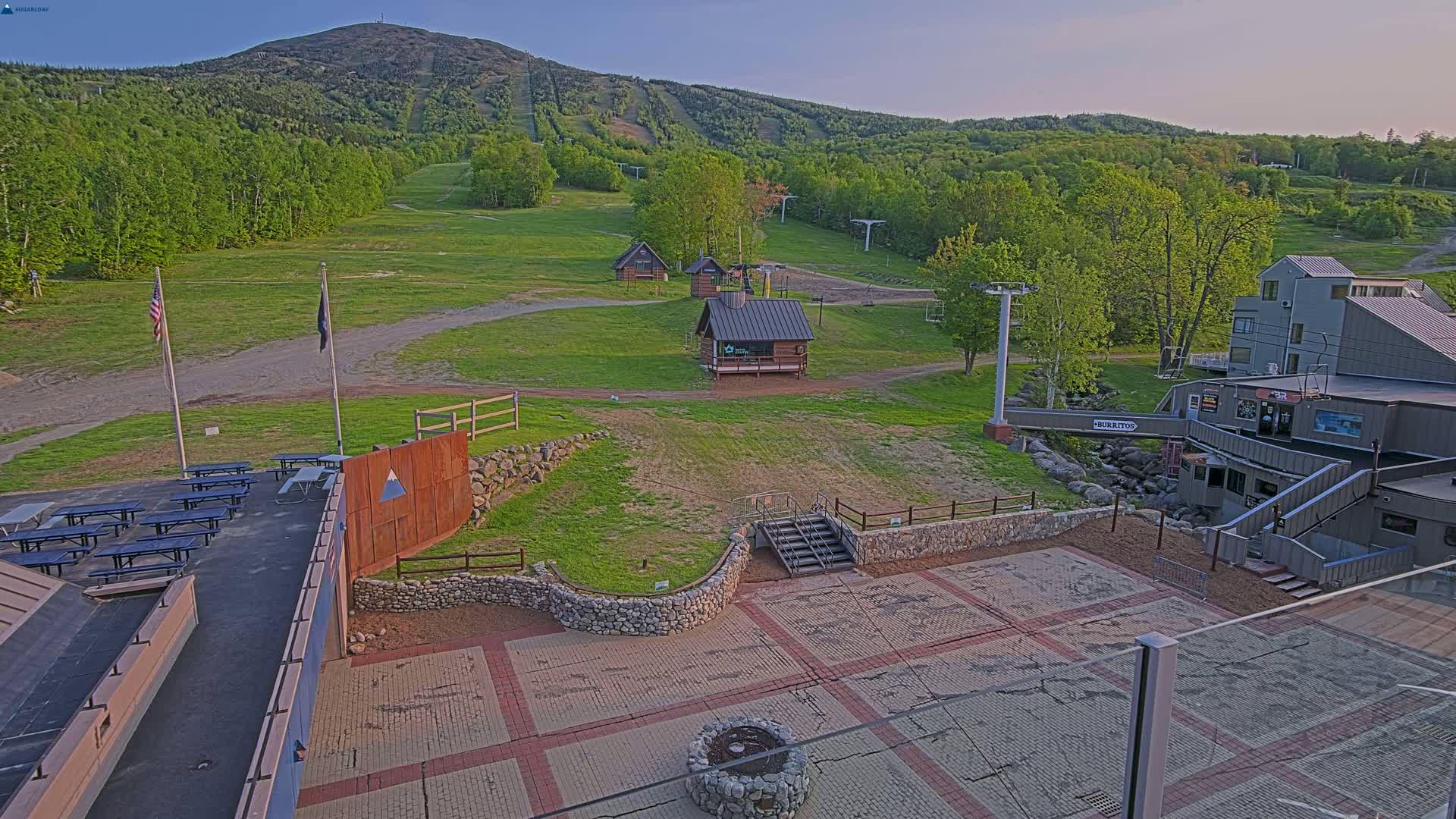 An outdoor scene at a ski resort shows a paved area with picnic tables, small buildings, a mountain in the background, and green, leafy trees under a clear sky.