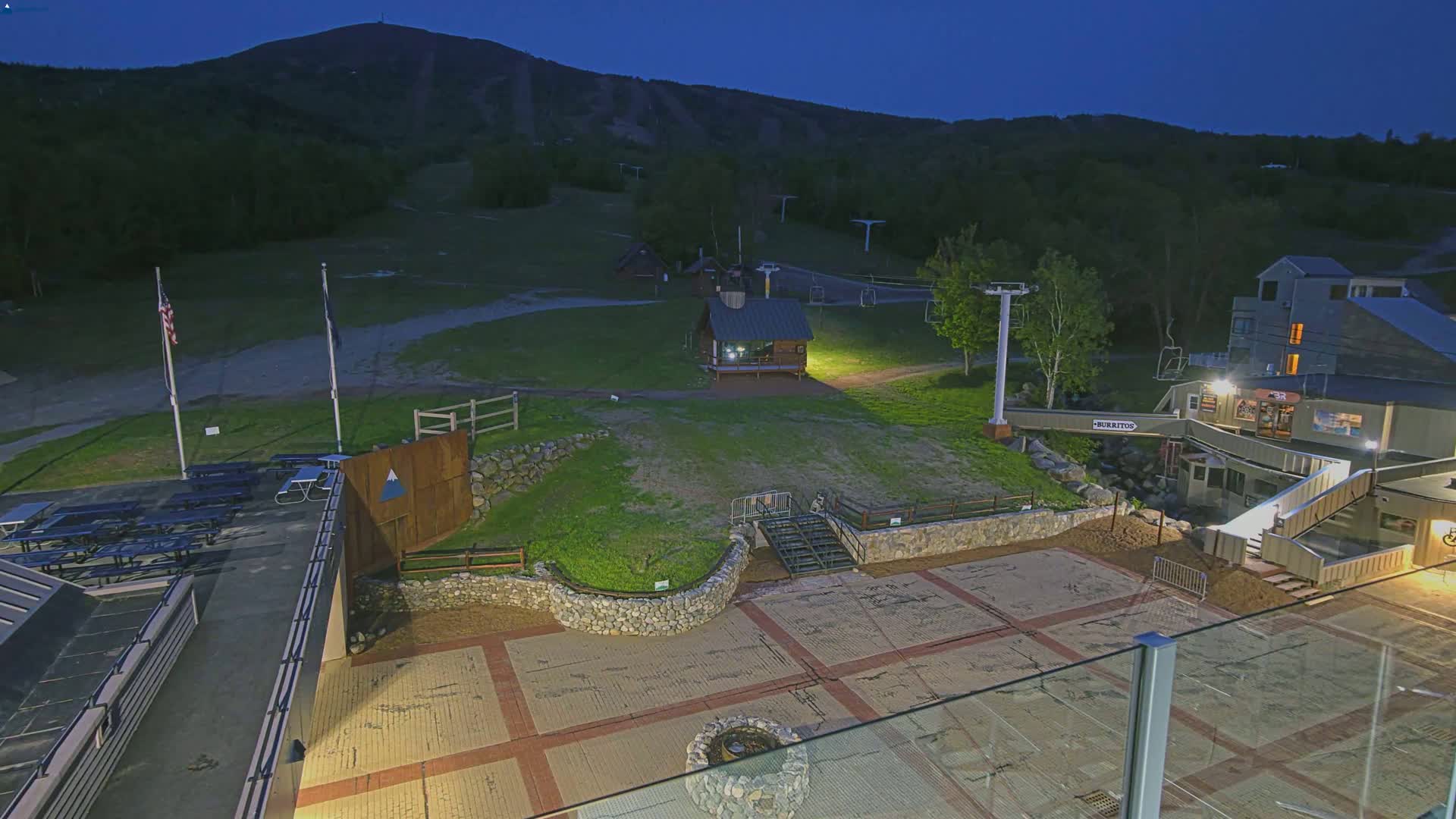 A nighttime view of a ski resort base area with picnic tables, a small building, a ski lift, and a paved plaza, all set against a dark mountain under a clear sky.
