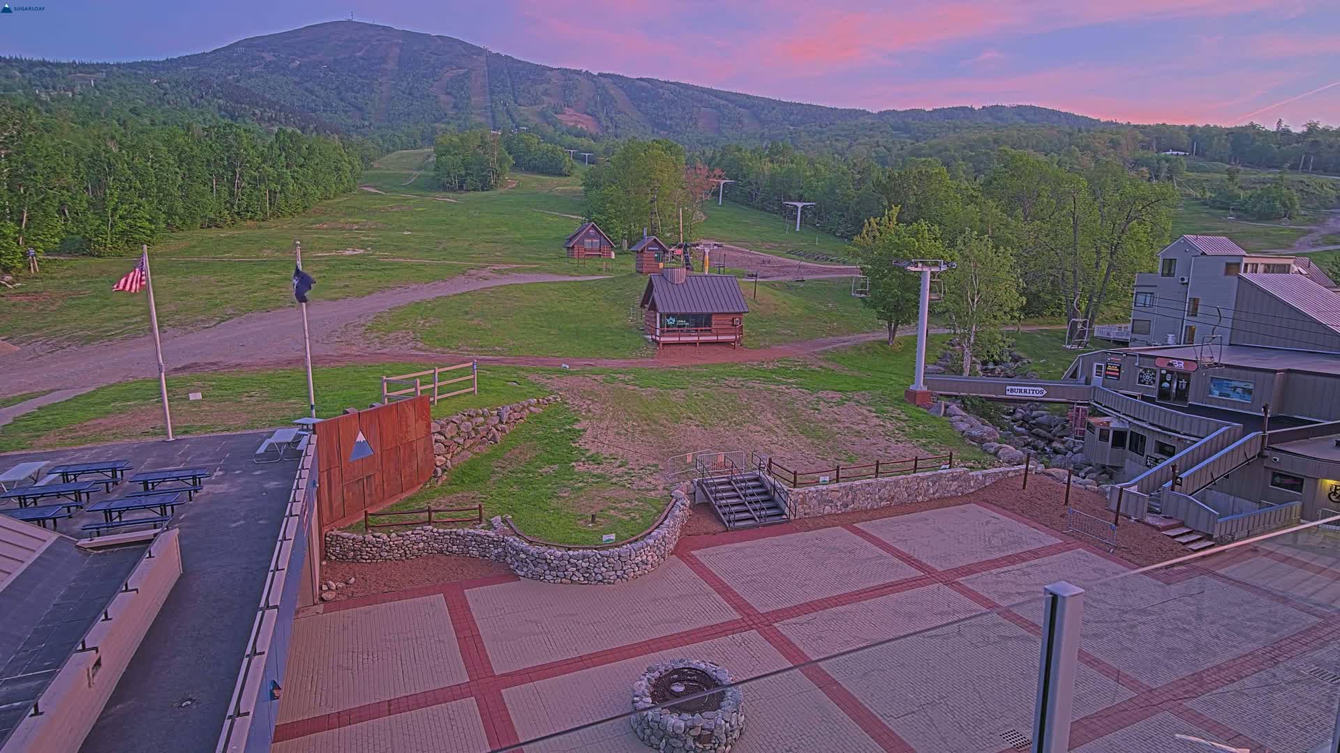 An outdoor paved area with picnic tables, small buildings, and a ski lift against a backdrop of a mountain at sunset.