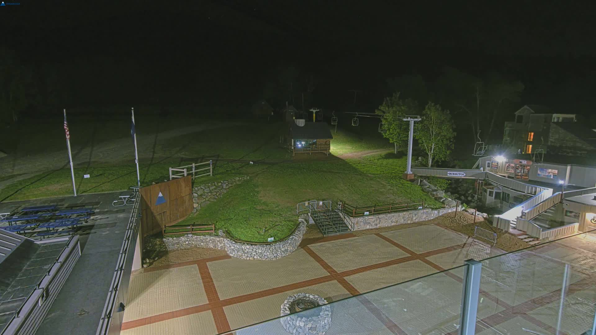 An illuminated outdoor area at night features a paved plaza, a small building, a ski lift, picnic tables, and a grassy hill.