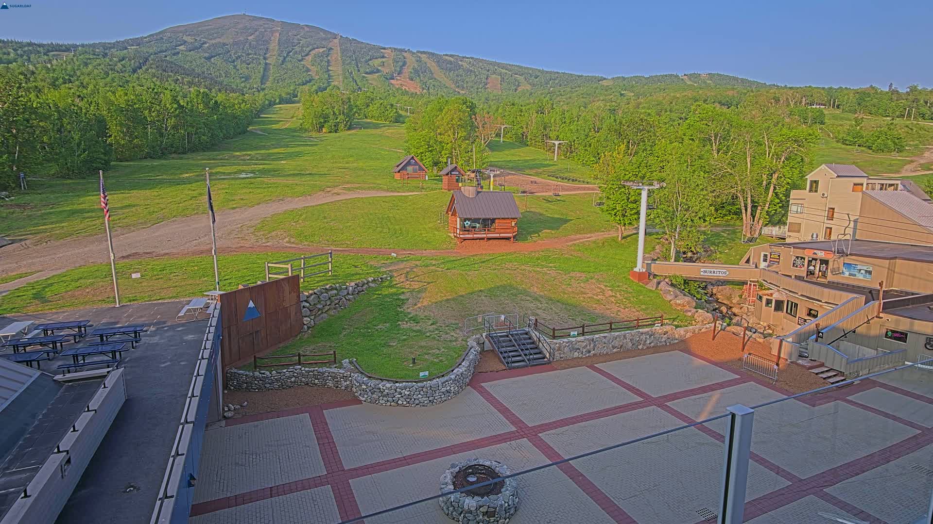 An outdoor scene of a ski resort in the summertime shows a grassy area with small cabins, a chairlift, and a paved patio area with picnic tables near a mountain under a clear blue sky.