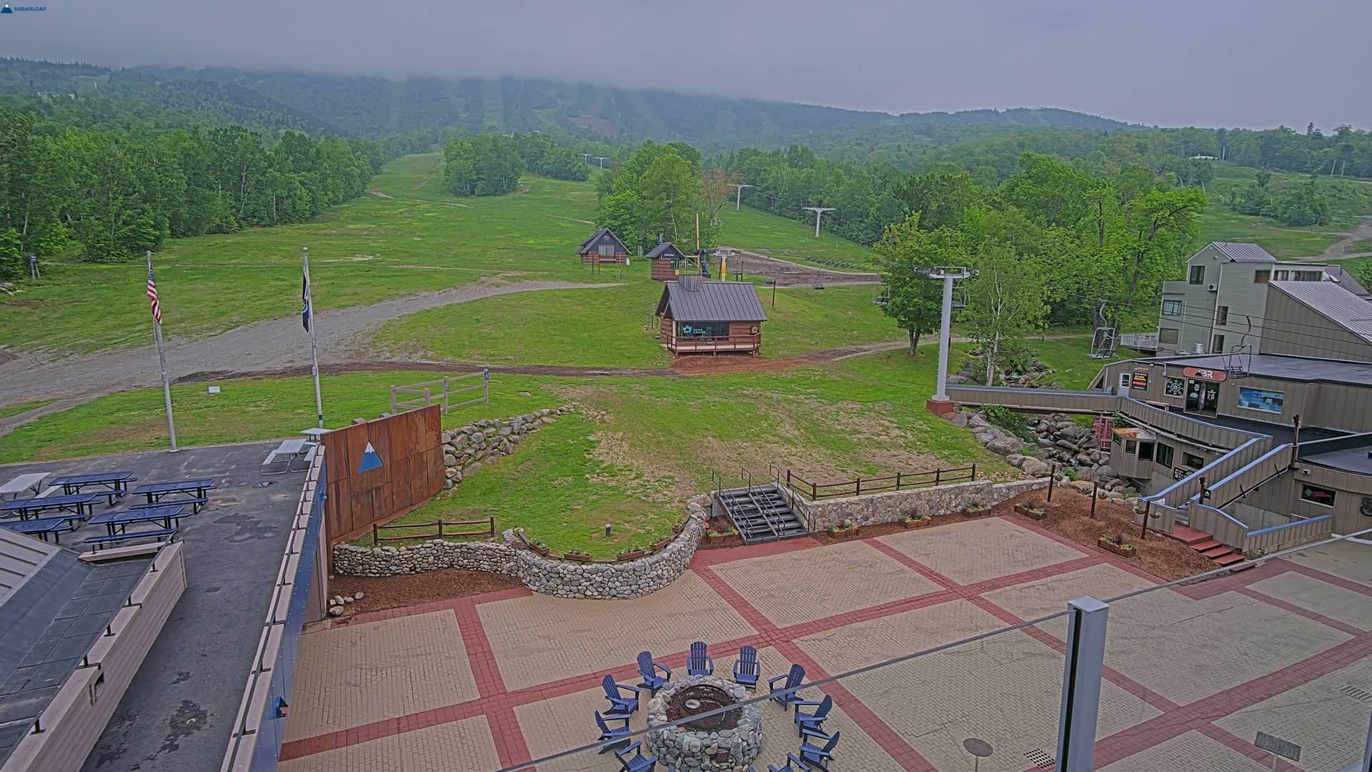 An outdoor patio area with picnic tables and a fire pit overlooks a grassy ski slope and mountain range under an overcast sky.
