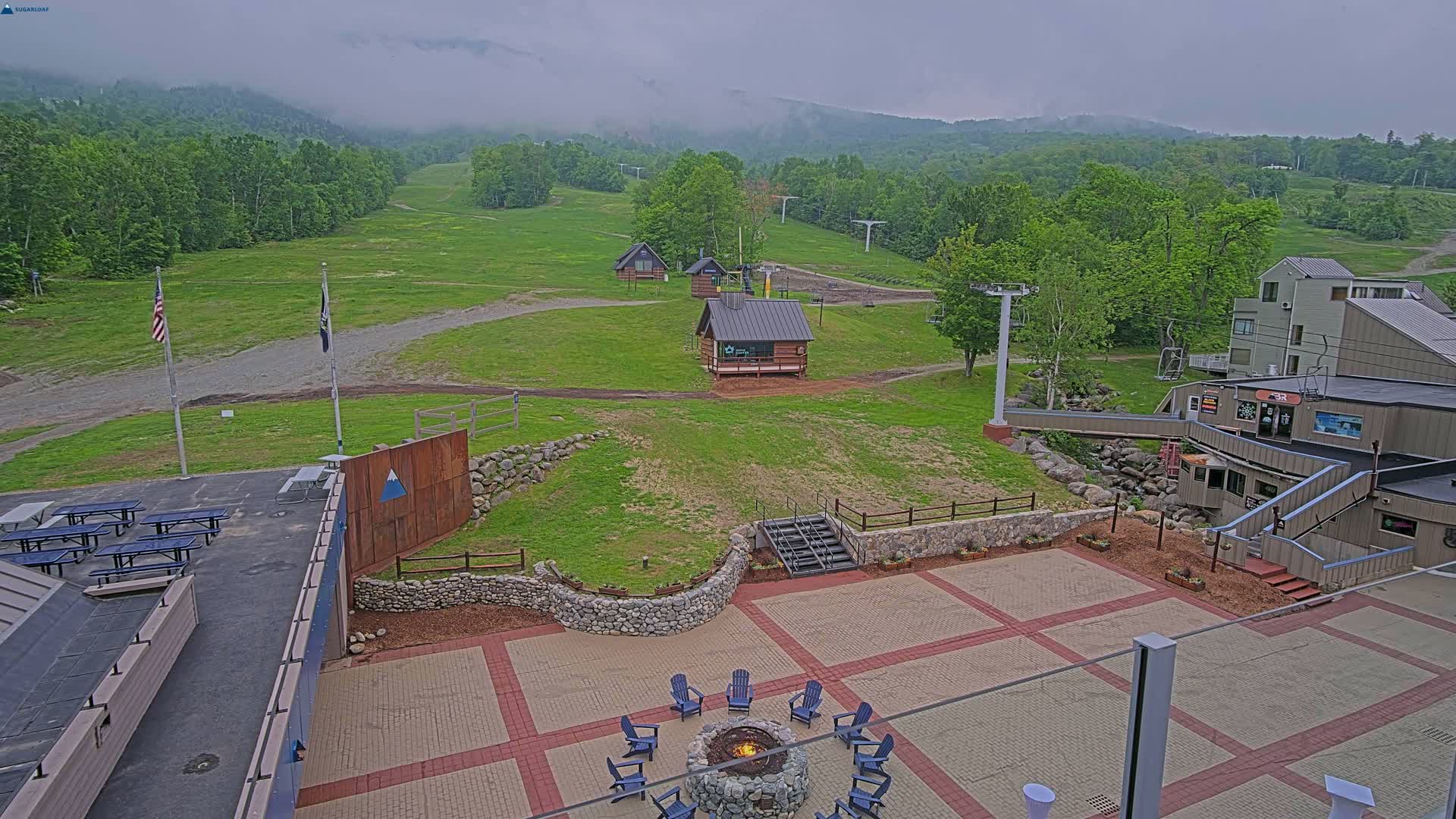 An outdoor patio area with a fire pit and seating, overlooking grassy ski slopes and mountains shrouded in fog.