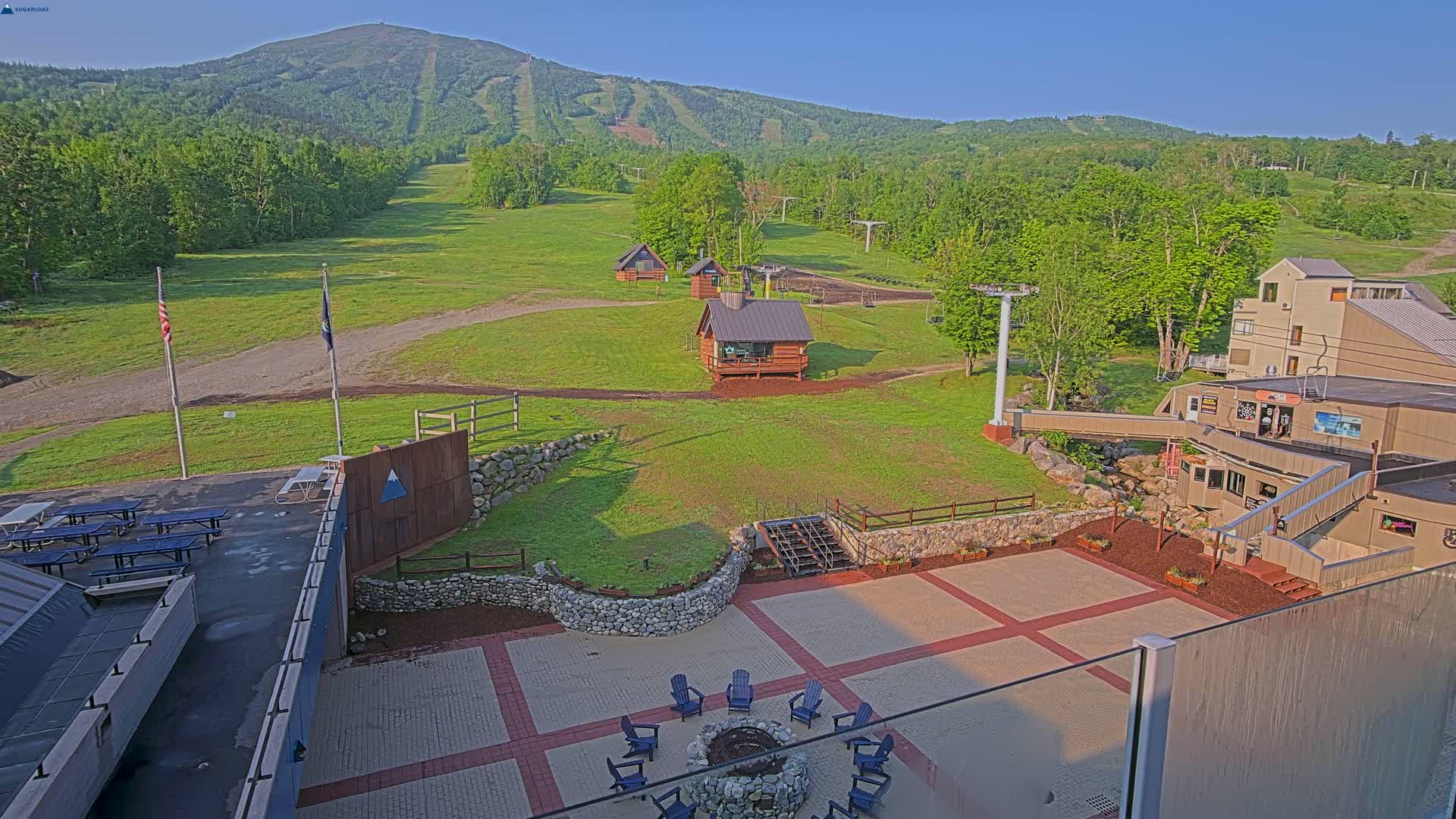 An outdoor patio area with a fire pit overlooks a grassy ski slope and mountain under a clear, sunny sky.