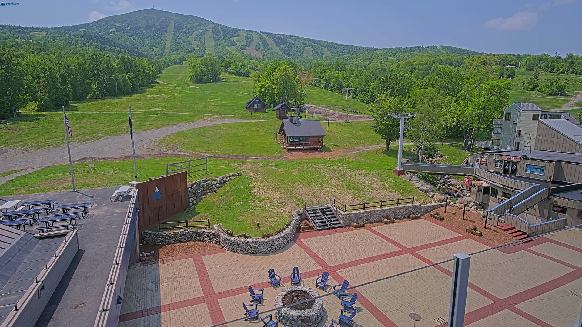 A sunny outdoor view of a ski resort in the summer, showing a grassy hillside with ski lifts, small buildings, picnic tables, and a fire pit surrounded by blue chairs.