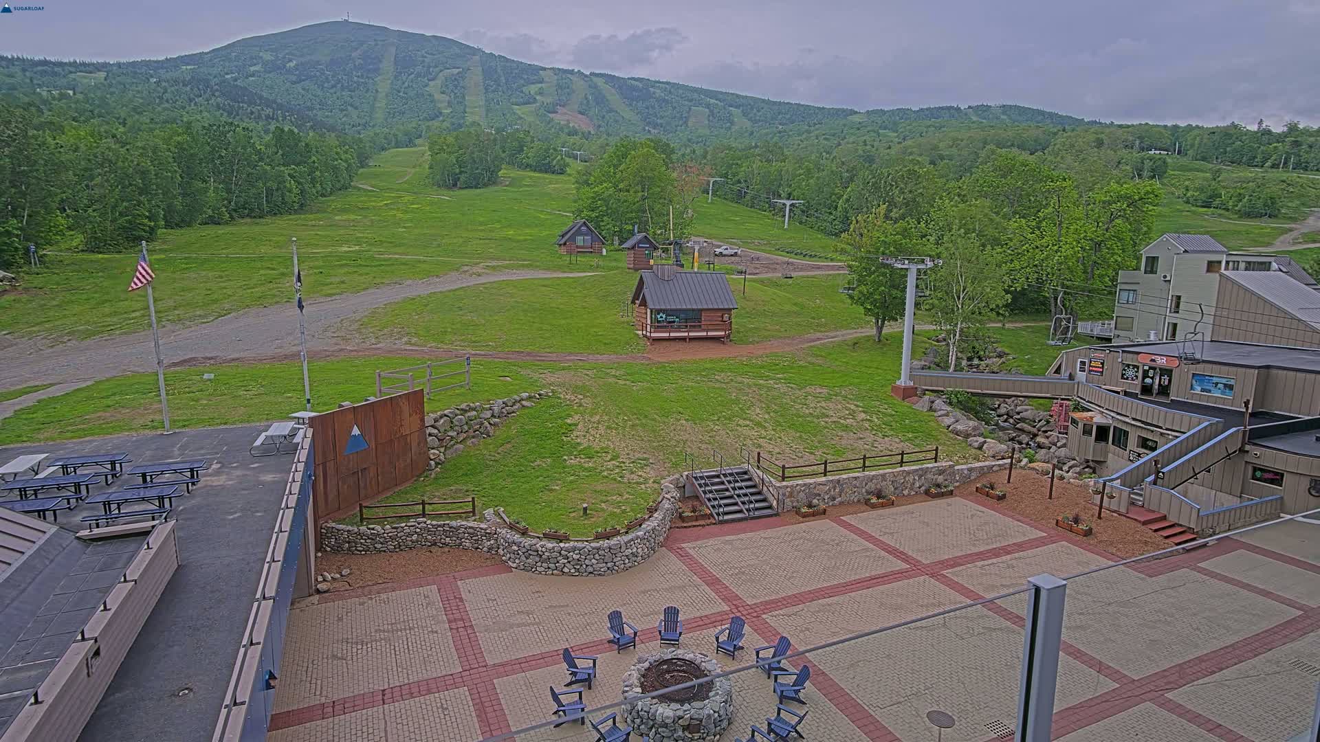 An outdoor patio area with a fire pit and seating, overlooking a grassy ski slope and mountain under an overcast sky.