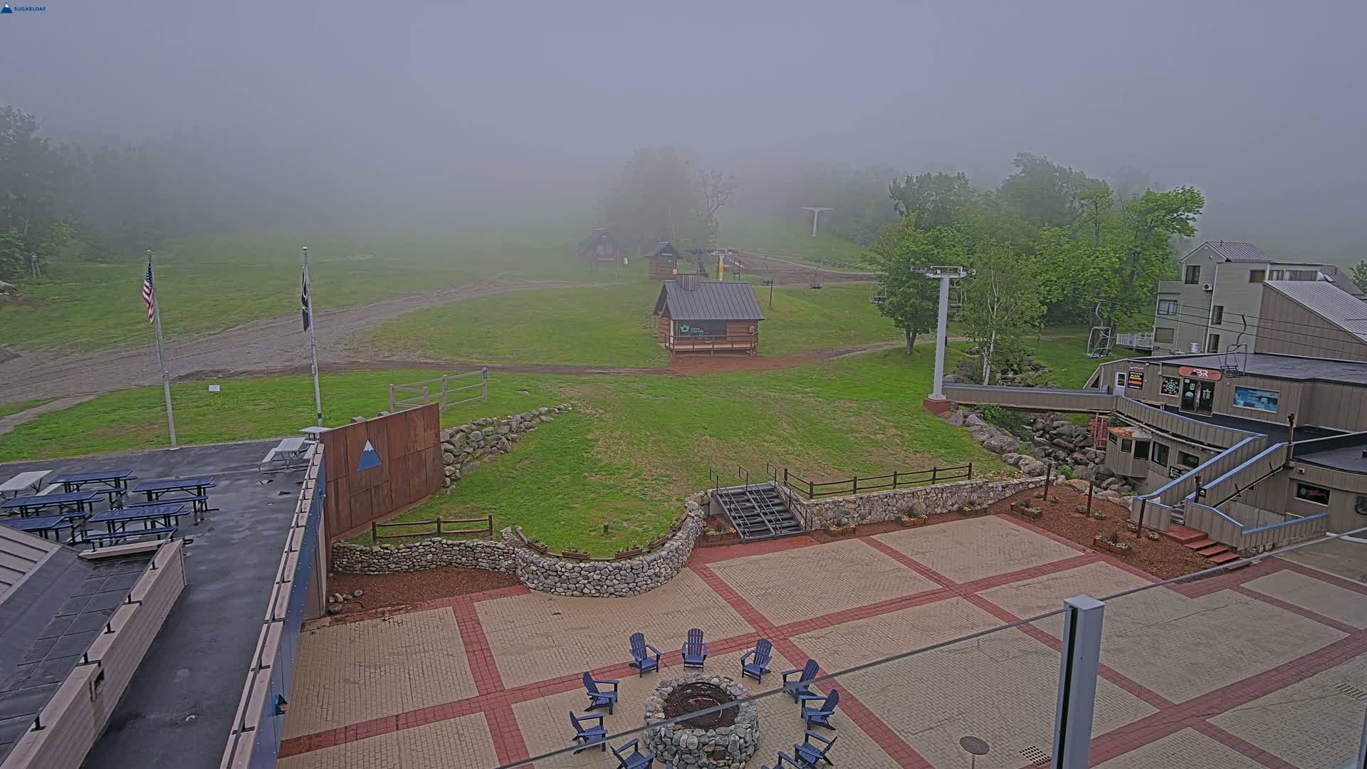 A foggy view of a ski resort's base area shows several small buildings, a fire pit with chairs, picnic tables, and a chairlift in the distance.