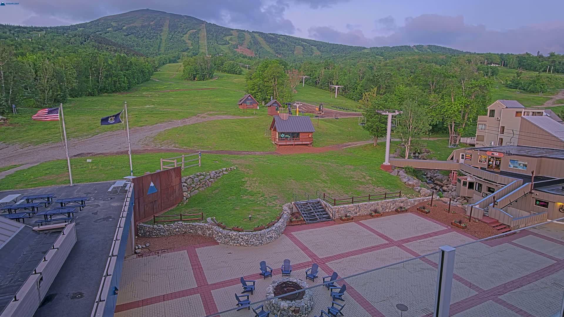 An outdoor patio area with a fire pit and chairs overlooks a grassy ski slope and mountain under a partly cloudy sky.