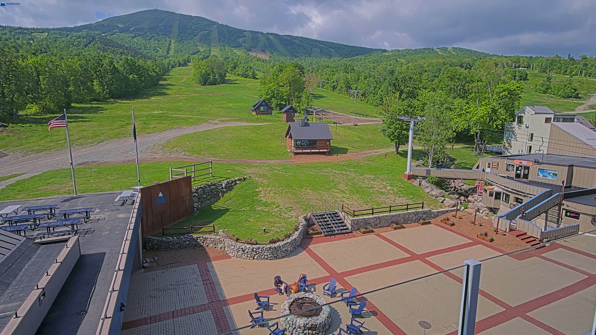 A paved outdoor area with picnic tables, a fire pit, and small buildings sits below a mountain with ski lifts and green slopes under partly cloudy skies.