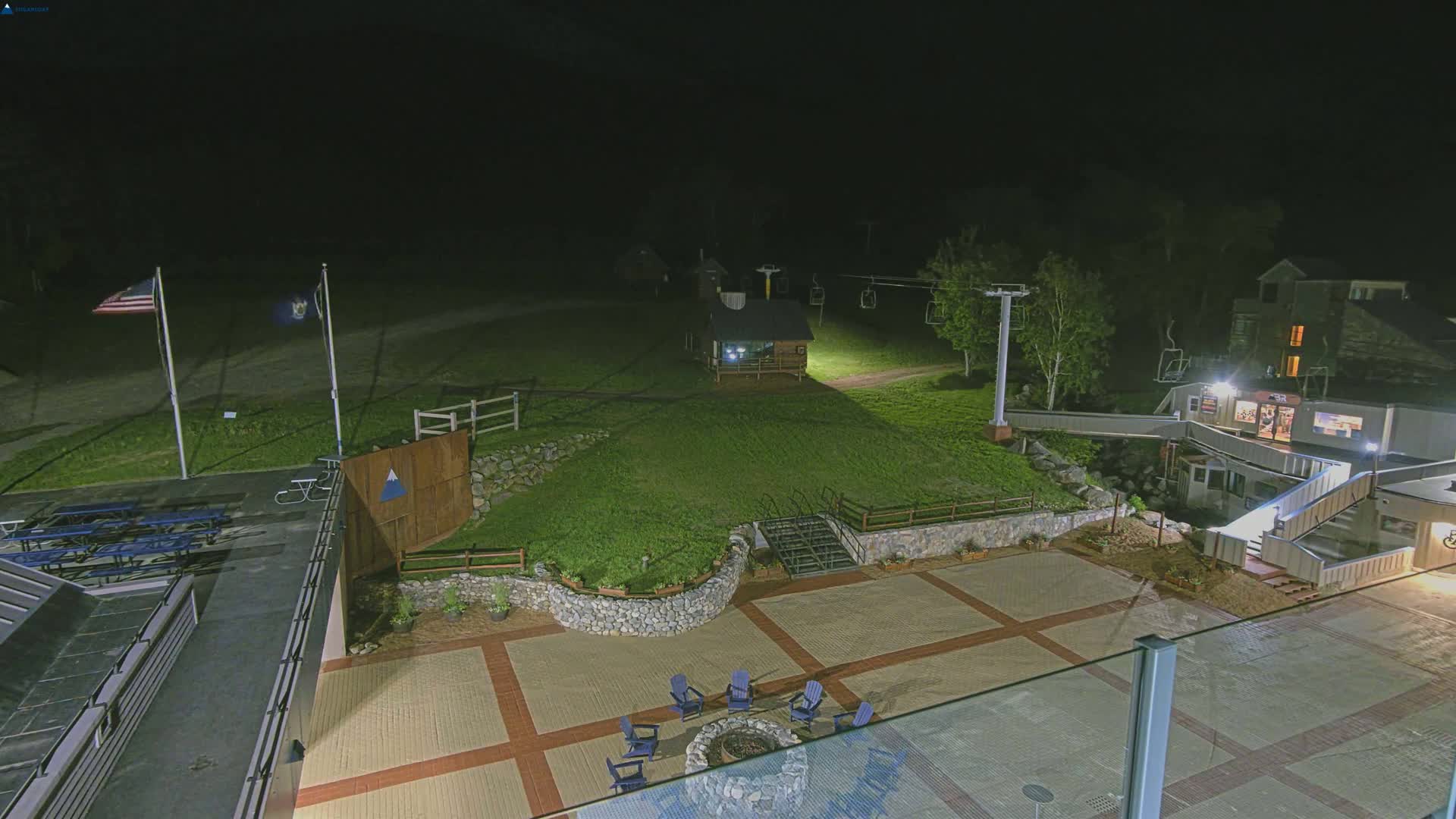 A nighttime aerial view of a paved outdoor area with a fire pit, seating, and a ski lift in the background under a dark sky.