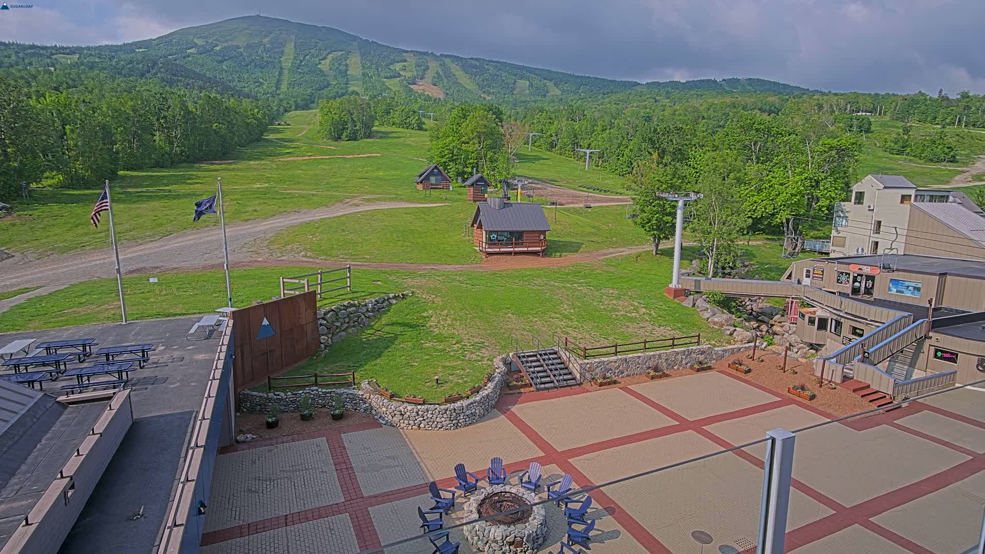 An outdoor patio area with picnic tables and a fire pit overlooks a grassy ski resort with several small buildings and a mountain in the background under partly cloudy skies.