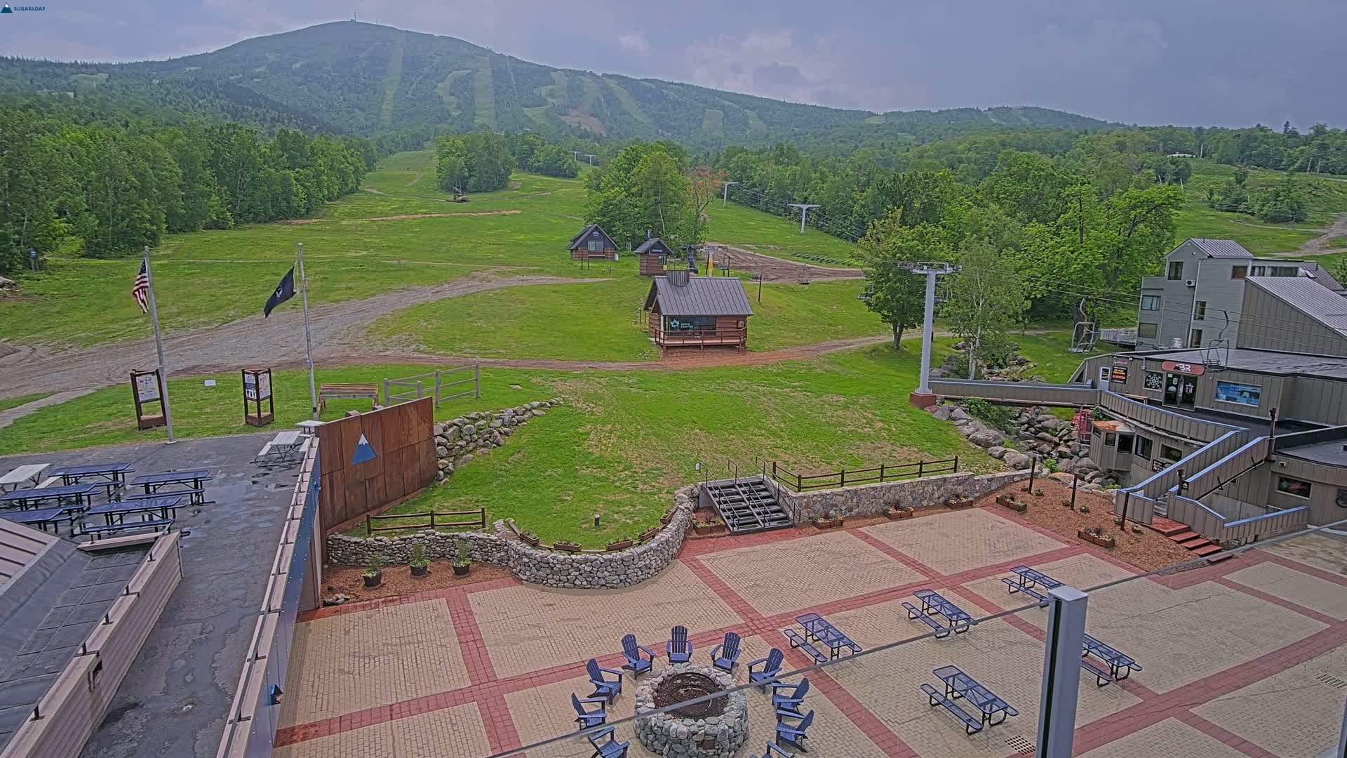 An outdoor patio area with picnic tables and a fire pit overlooks a grassy ski resort with a mountain in the background under overcast skies.