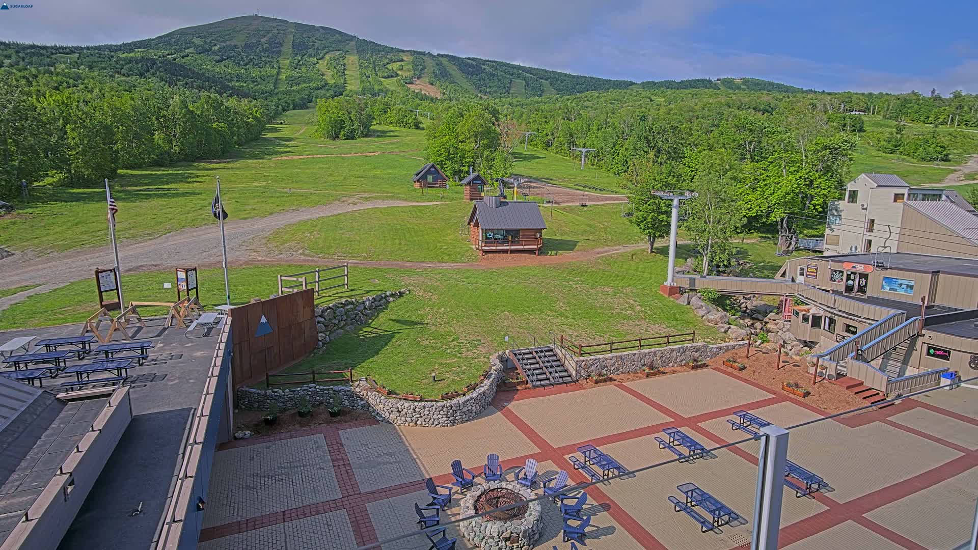 A mountain ski resort in the summertime shows grassy slopes, small buildings, picnic tables, a fire pit, and a paved patio area under a mostly clear sky.