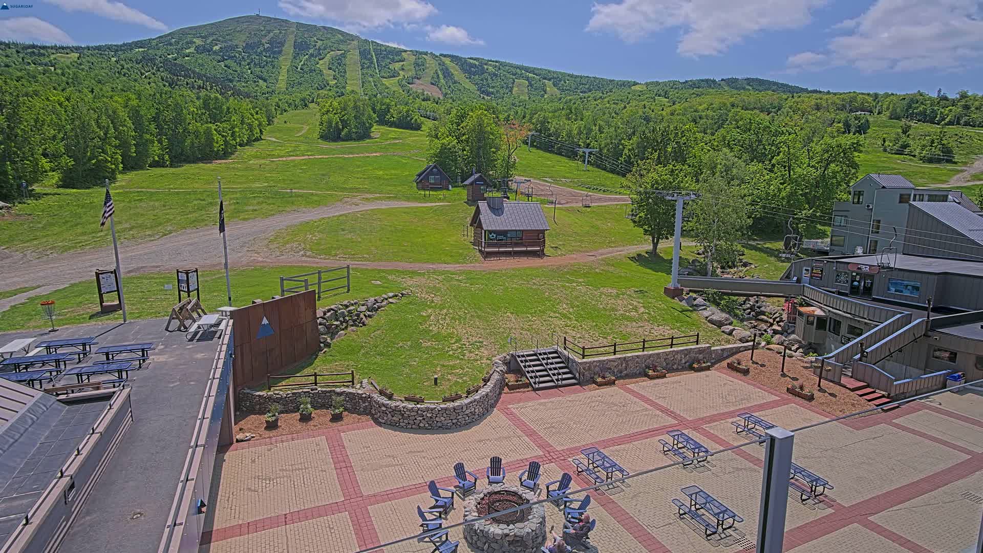 An outdoor patio area with picnic tables and a fire pit overlooks a grassy hillside with ski lift infrastructure and a mountain in the background under sunny skies.