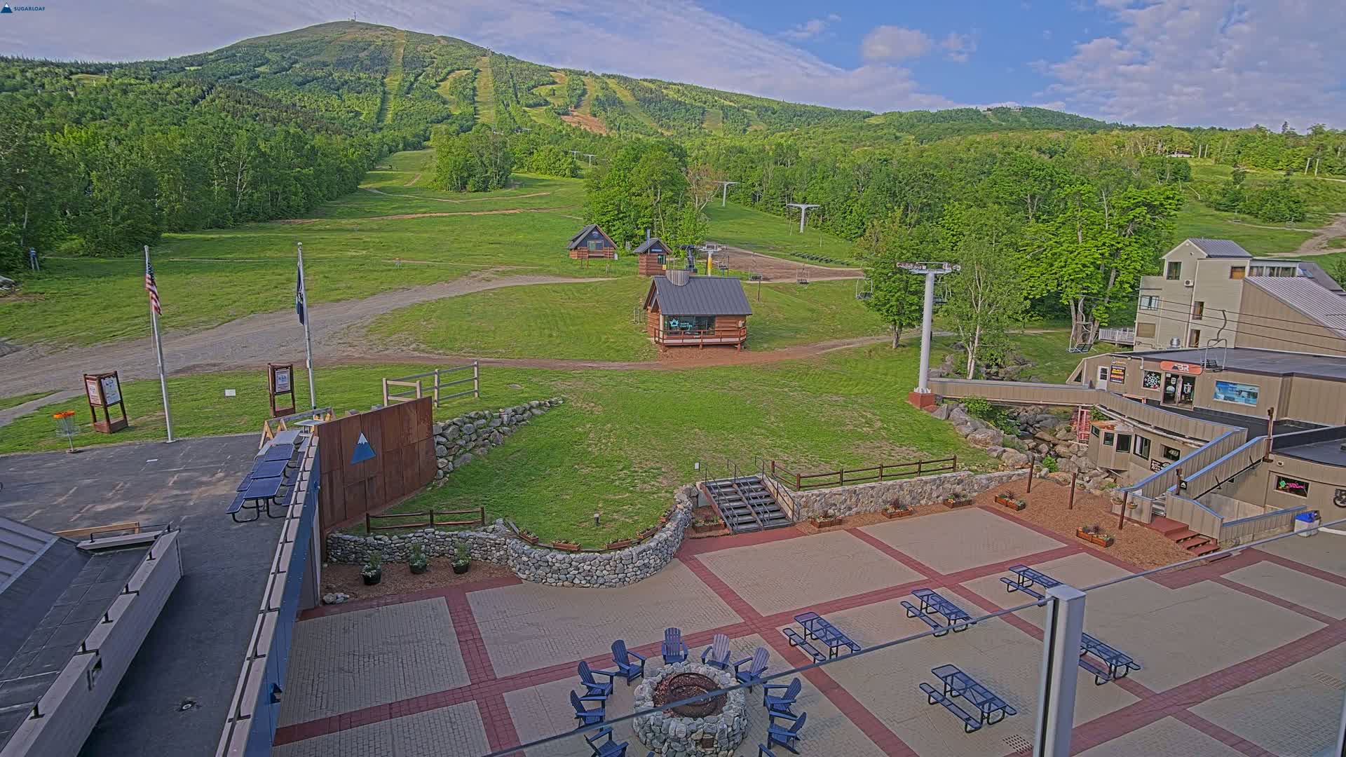 An outdoor patio area with picnic tables and a fire pit is situated below a ski resort, overlooking a grassy mountainside under a mostly clear sky.