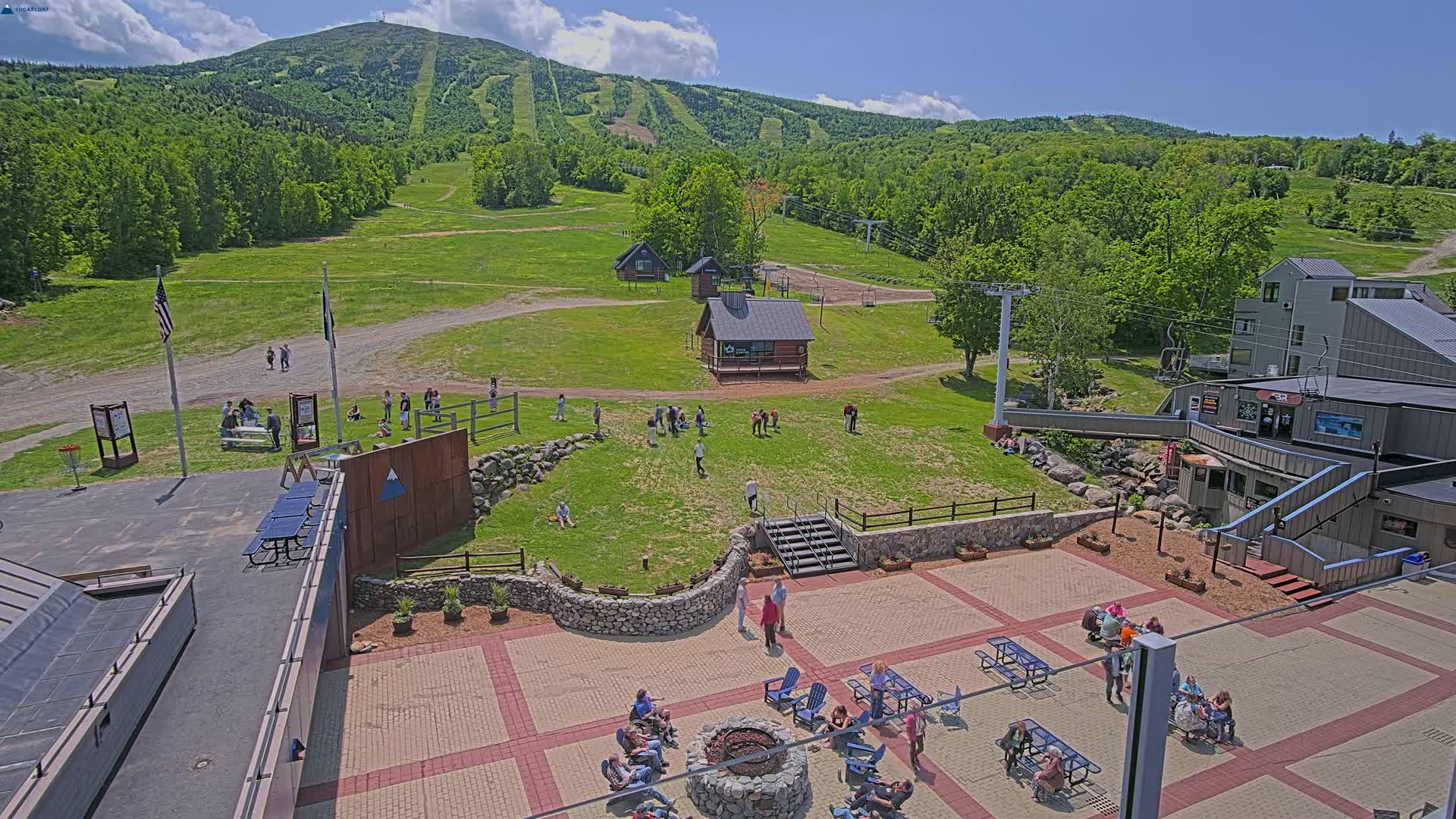 An outdoor patio area with people relaxing and socializing, situated on a grassy hillside overlooking a mountain with ski trails, under a partly cloudy sky.