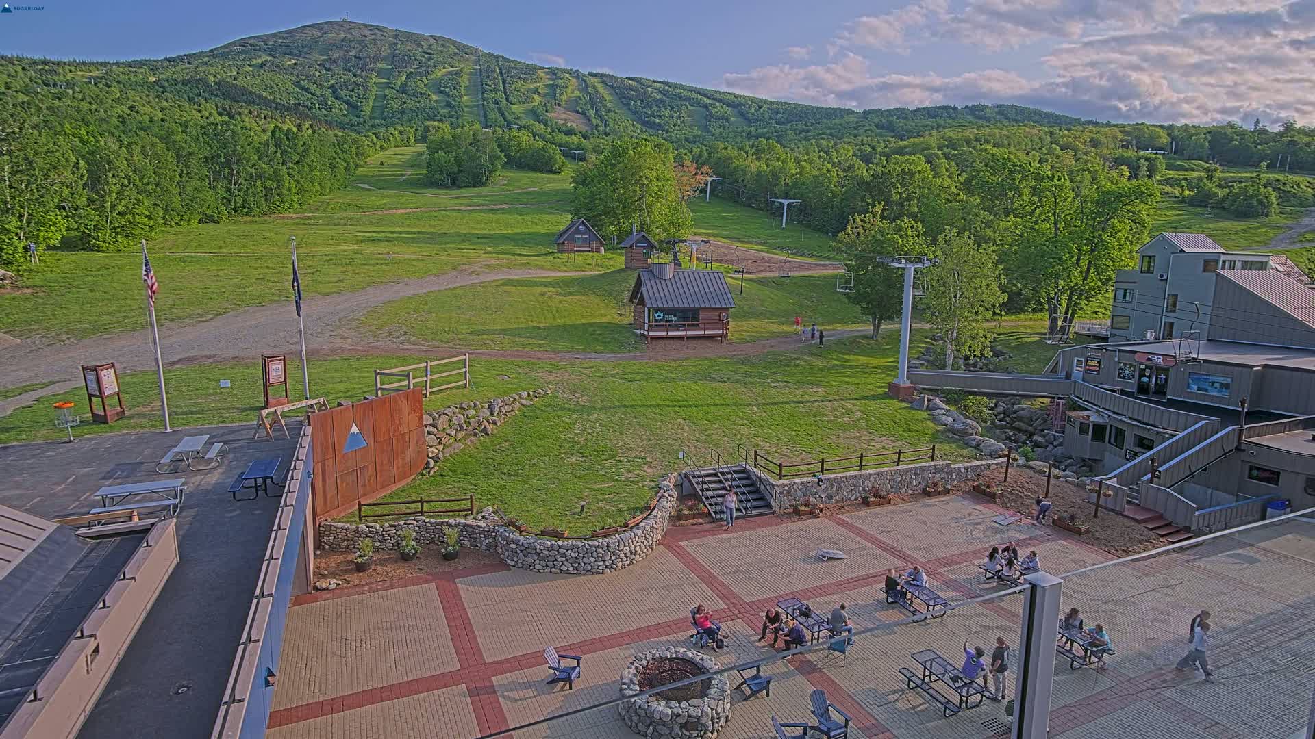 An outdoor patio area with people relaxing and socializing, situated on a grassy hillside overlooking a mountain with ski trails, under a partly cloudy sky.
