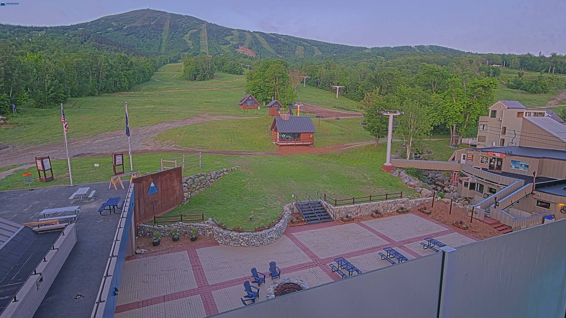 An outdoor patio area with picnic tables and a fire pit overlooks a grassy ski resort with several small buildings and a mountain in the background under a twilight sky.