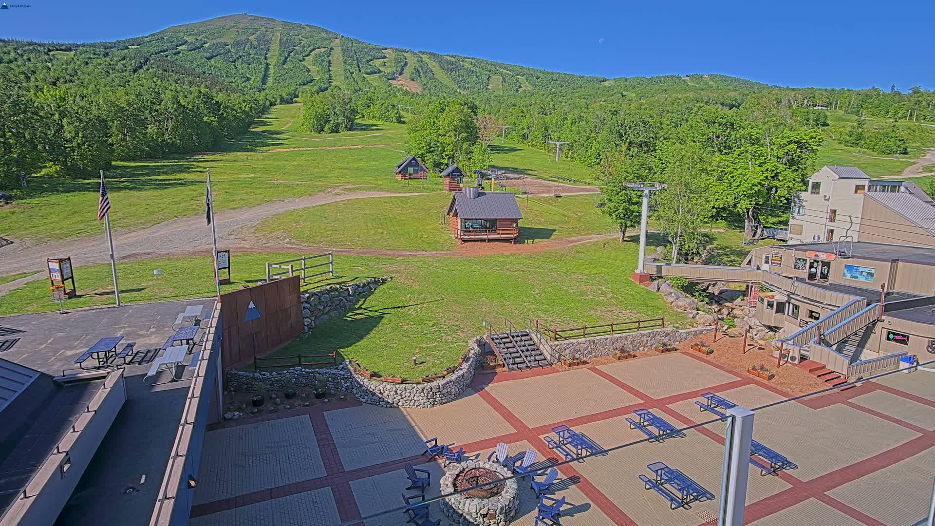 An outdoor patio area with picnic tables and a fire pit overlooks a grassy ski resort with several small buildings and a mountain in the background under sunny skies.