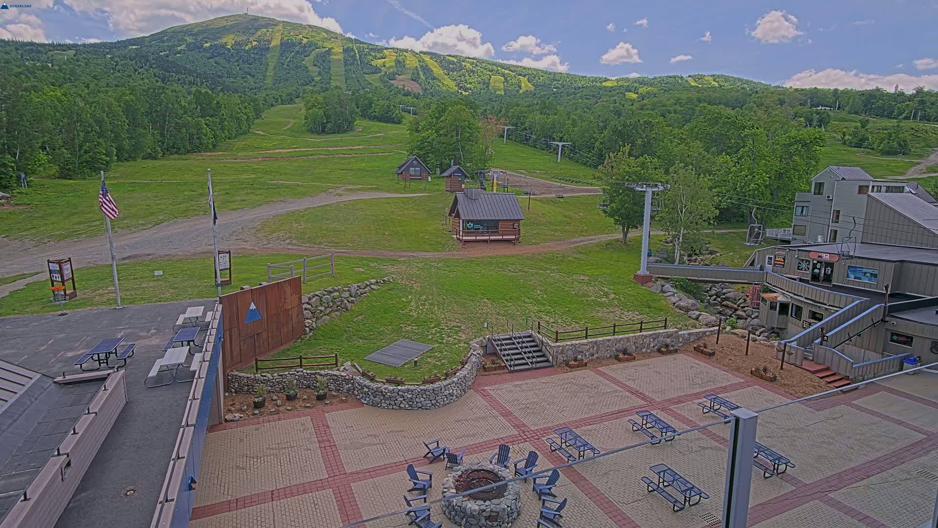 A paved outdoor patio area with picnic tables and a fire pit is situated below a ski resort with several small buildings and chairlifts, all set against a backdrop of a lush green mountain under a partly cloudy sky.