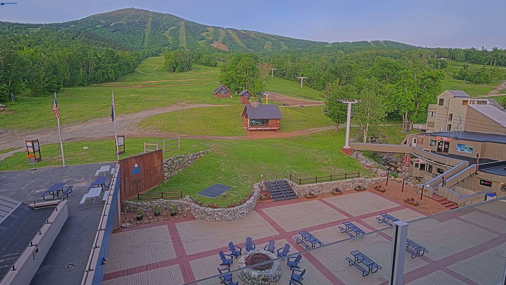 An outdoor patio area with picnic tables and a fire pit is situated below a ski resort, overlooking a grassy landscape and a mountain under a clear sky.