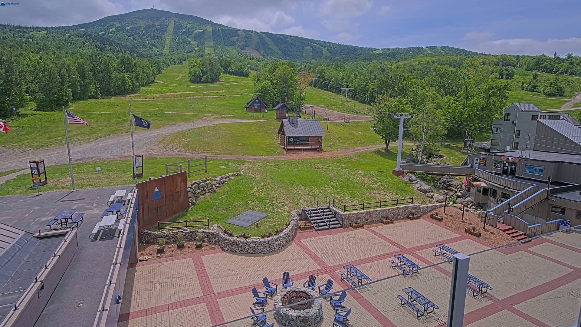 An outdoor patio area with picnic tables and a fire pit is situated below a ski resort with several small buildings and a mountain in the background under partly cloudy skies.
