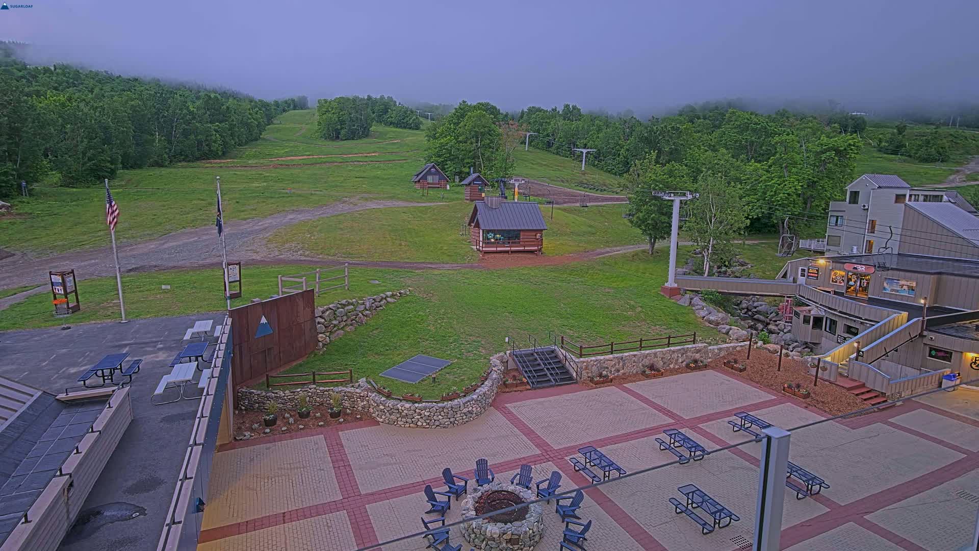 A paved patio area with picnic tables and a fire pit overlooks a grassy ski slope with lift equipment and small buildings under a foggy sky.