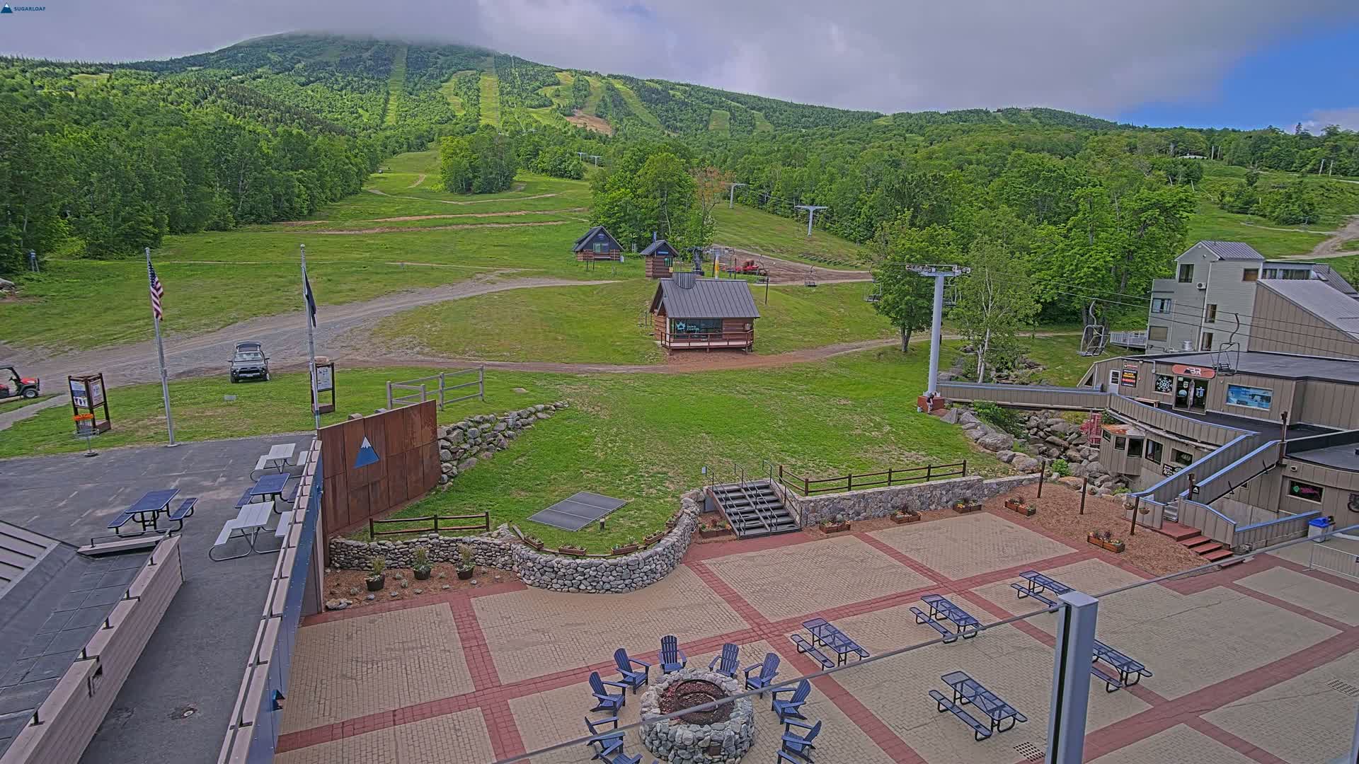 An outdoor patio area with picnic tables and a fire pit overlooks a grassy ski resort with several small buildings and chairlifts under a mostly cloudy sky.