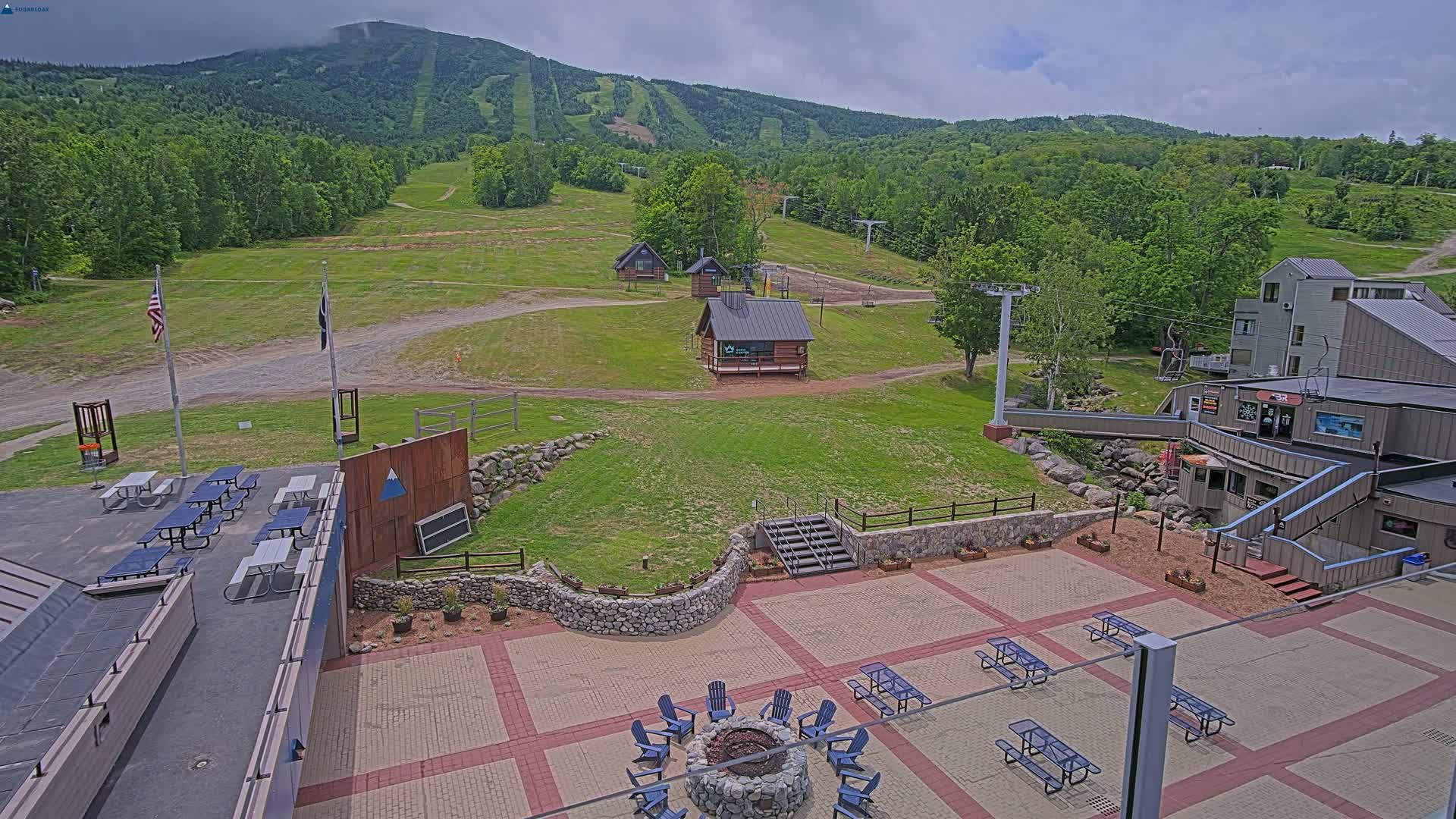 An outdoor patio area with picnic tables and a fire pit overlooks a grassy ski resort with several small buildings and a mountain in the background under partly cloudy skies.