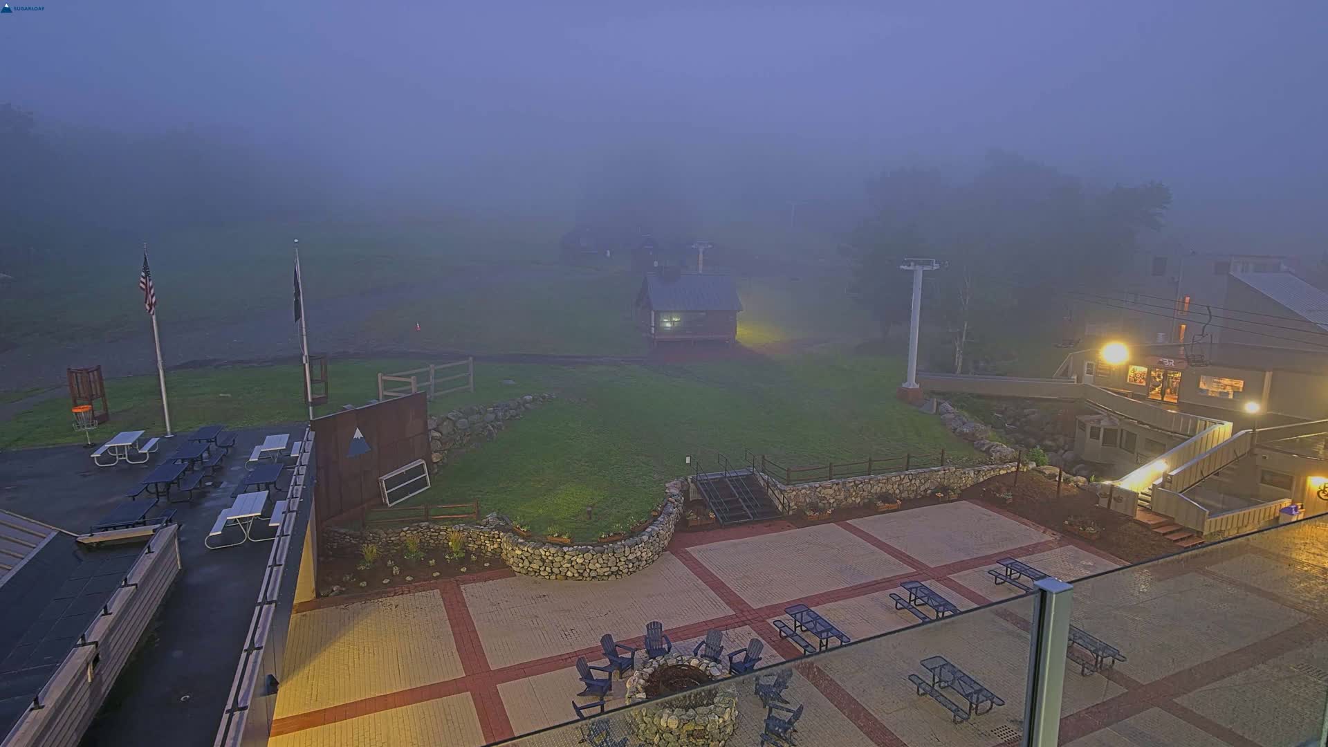 A foggy outdoor patio area with picnic tables, a fire pit, and a small building, overlooked by a building with a sloped roof.