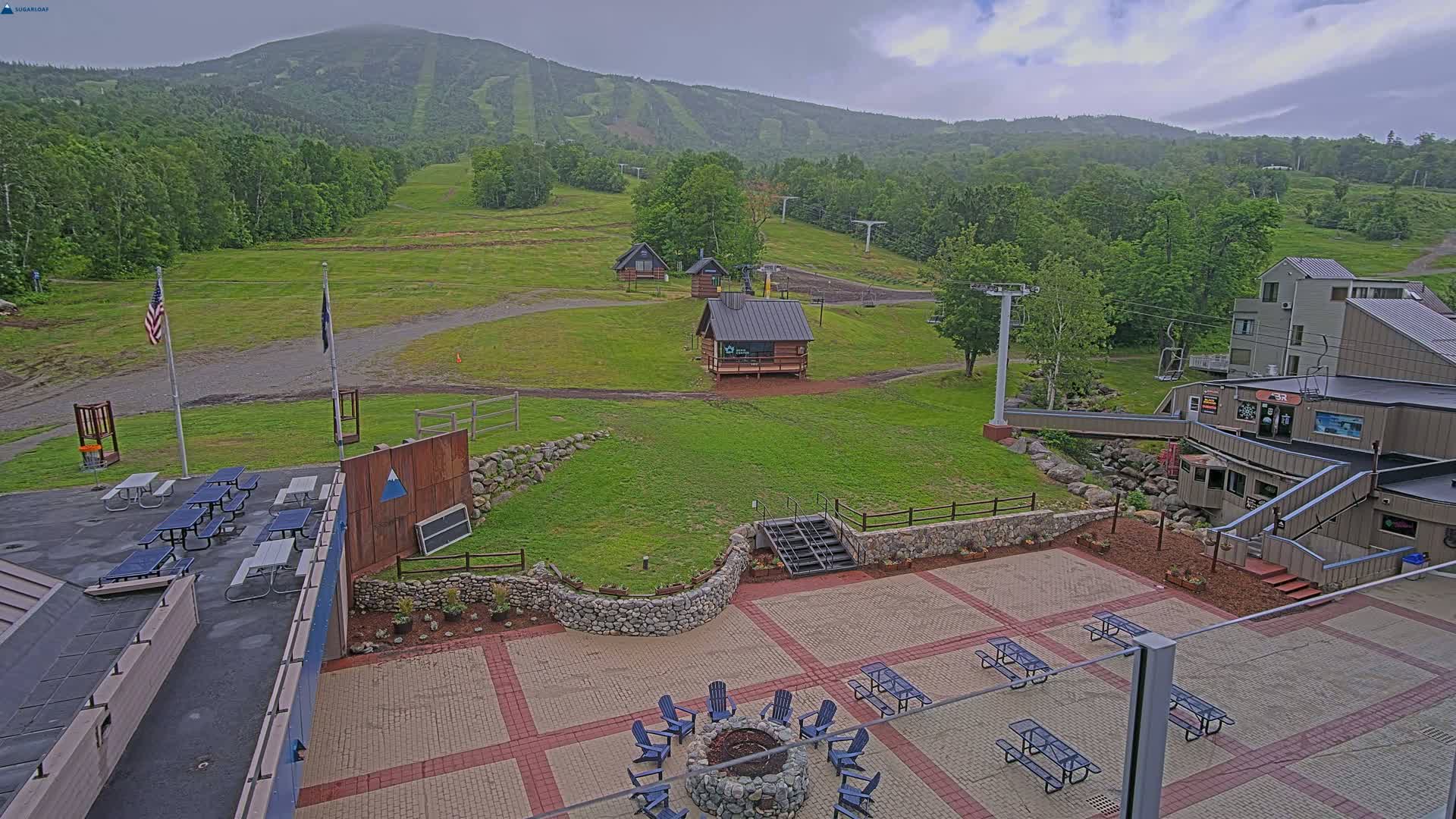 An outdoor patio area with picnic tables and a fire pit overlooks a grassy ski slope and mountain under cloudy skies.