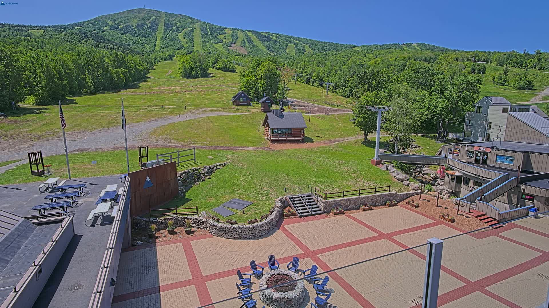A mountain ski resort with a paved patio area containing a fire pit and picnic tables, overlooks grassy slopes, ski lifts, and small buildings under a clear blue sky.