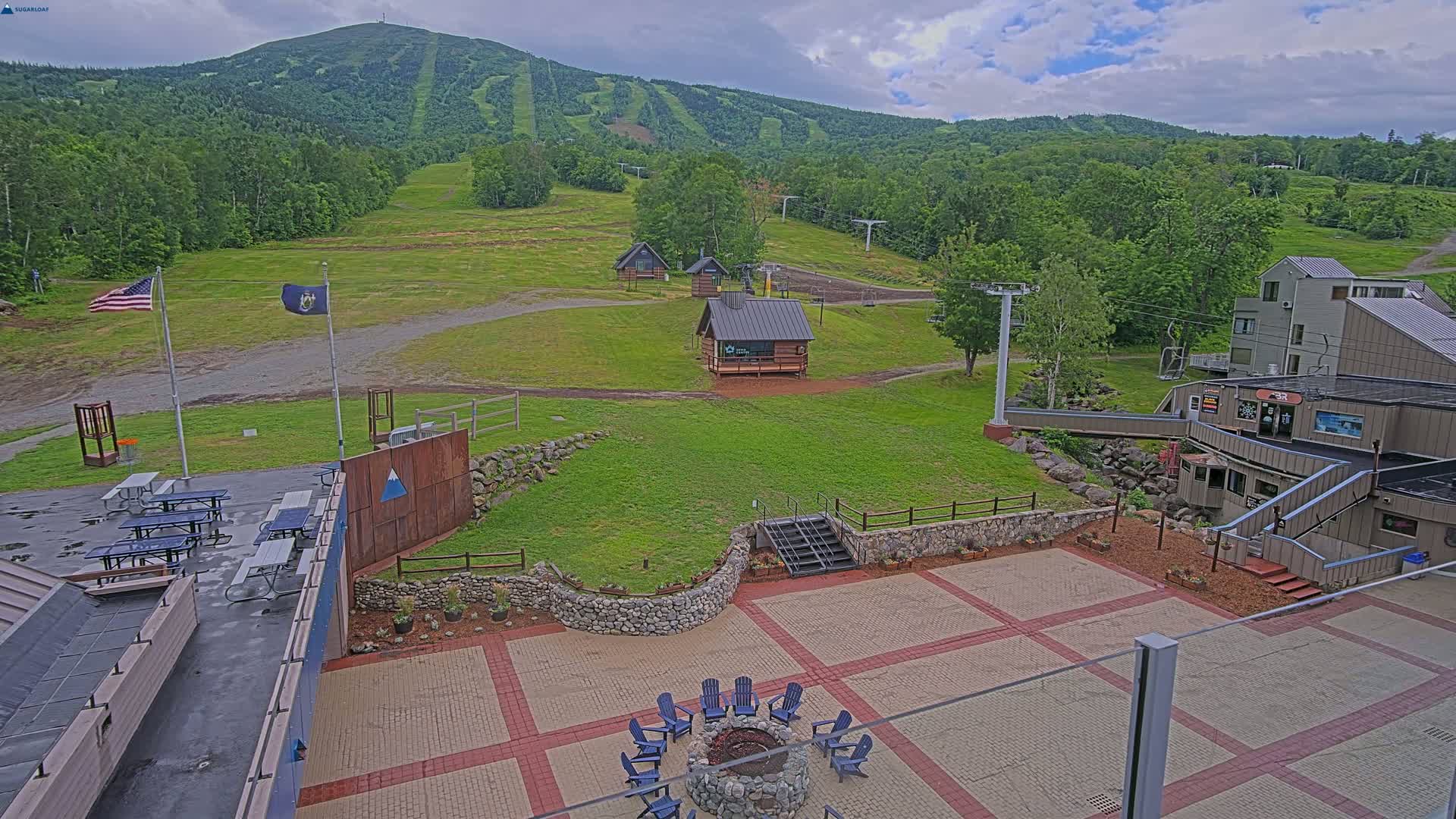 An outdoor patio area with picnic tables and a fire pit sits below a ski resort in front of a large mountain under a cloudy sky.