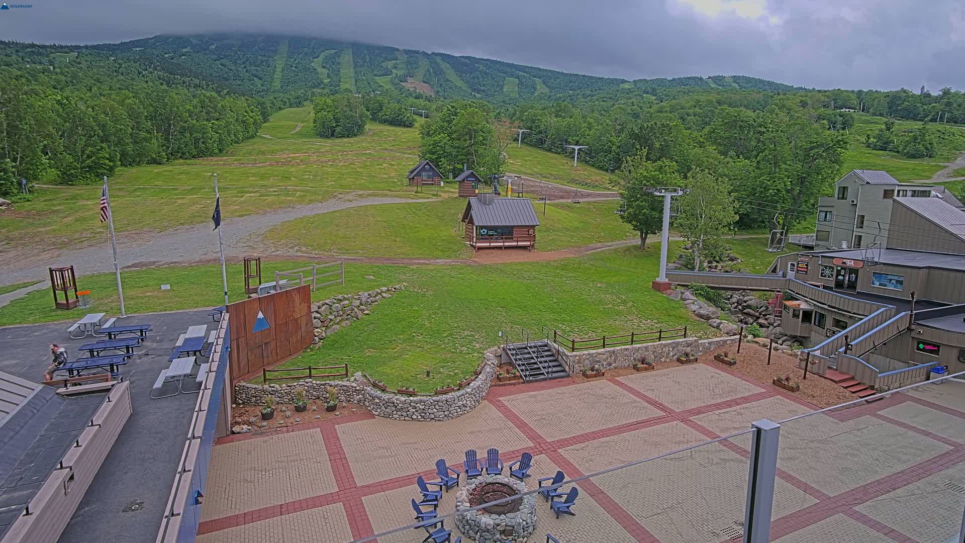 An outdoor patio area with picnic tables and a fire pit overlooks a grassy ski slope with ski lifts and several small buildings under an overcast sky.