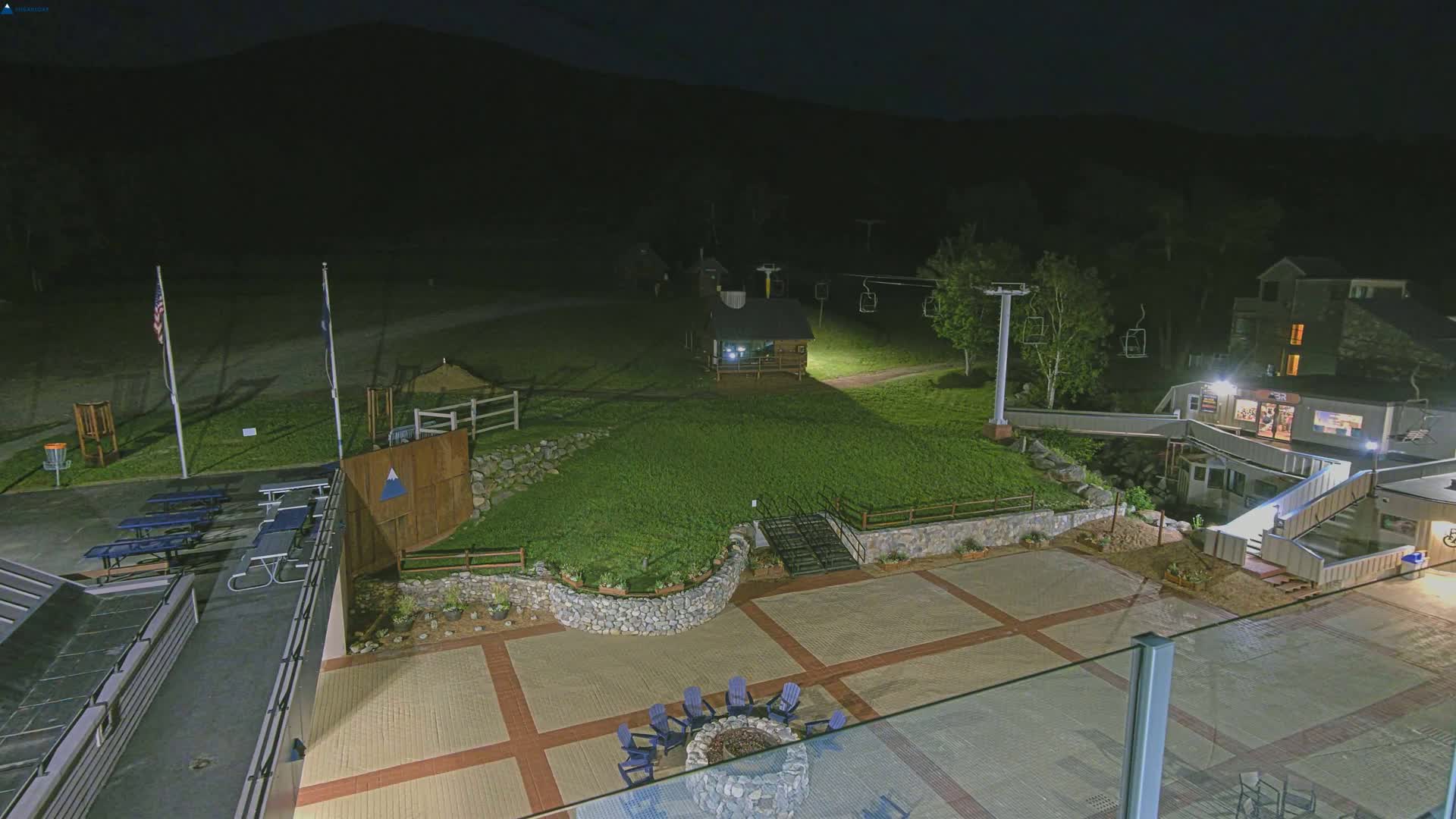 An outdoor nighttime scene at a ski resort shows a paved plaza with a fire pit and chairs, picnic tables, a ski lift, and a small building, all illuminated under dark skies.