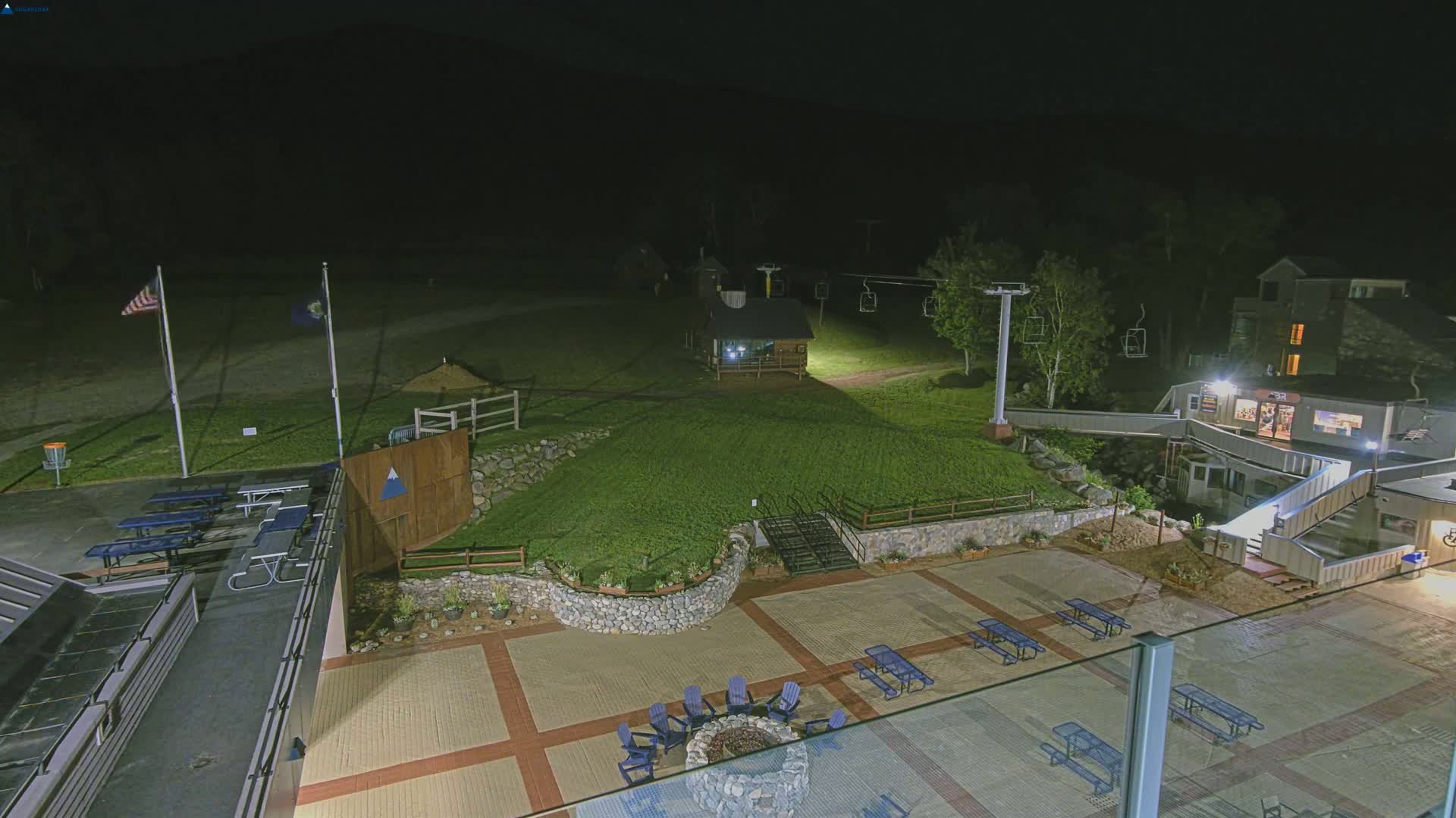 An outdoor nighttime scene shows a paved patio area with picnic tables and chairs surrounding a fire pit, adjacent to a grassy area with a ski lift and a small building, all illuminated by artificial light.