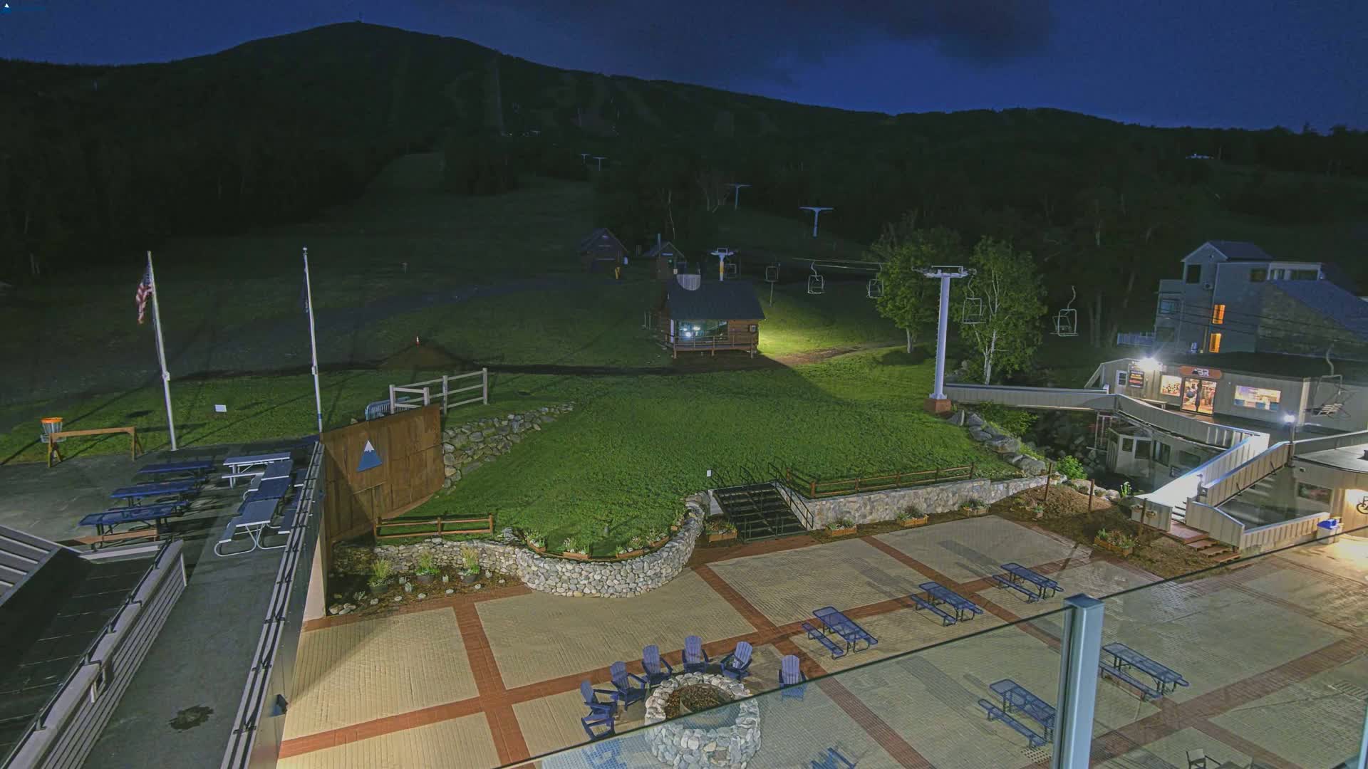 An outdoor nighttime scene shows a paved plaza with picnic tables and a fire pit, near ski lifts and a small building, all situated on a grassy hillside beneath a dark mountain.
