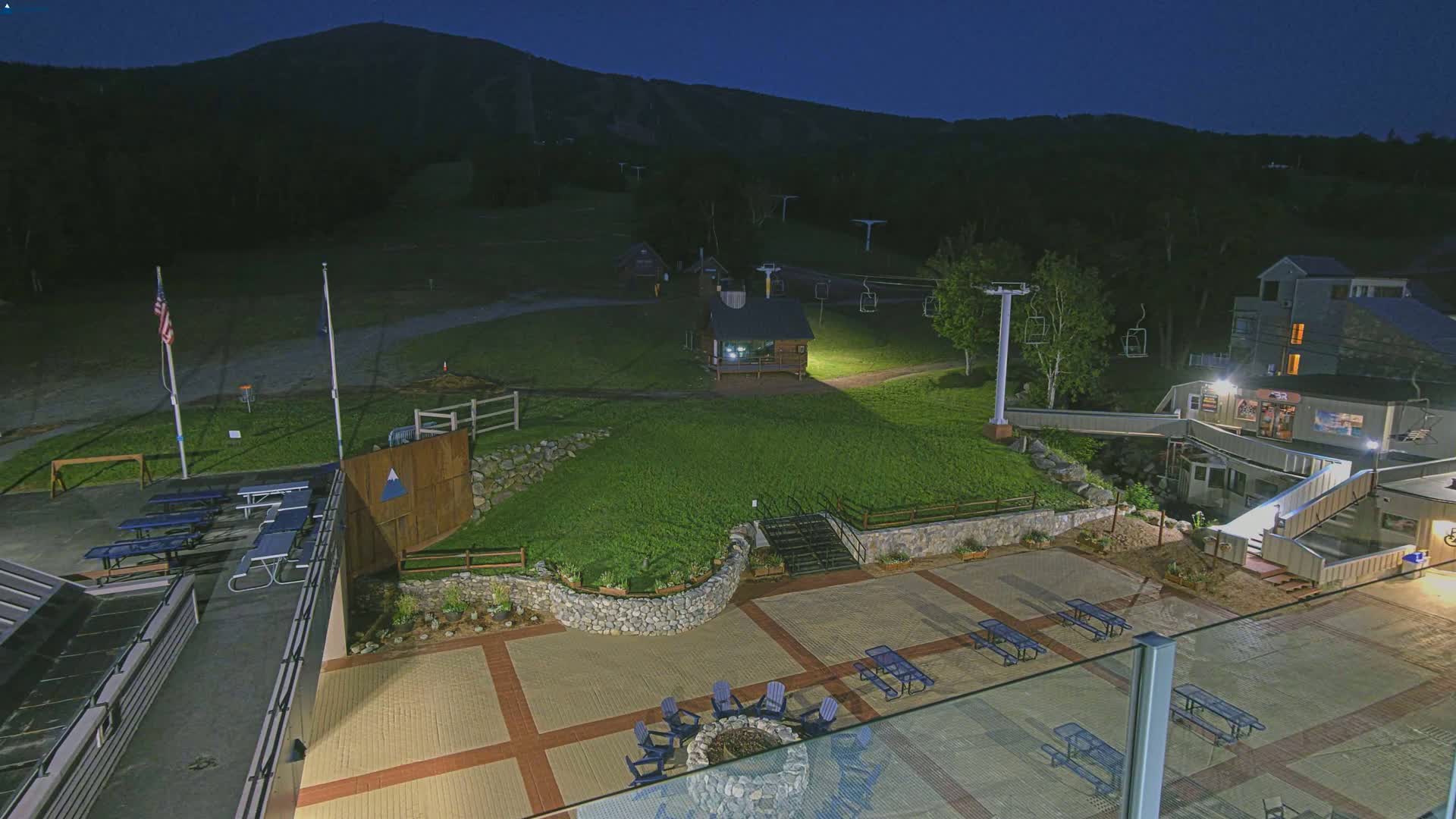 A nighttime view of a paved outdoor area with picnic tables, a fire pit, and a ski lift in the background under a clear, dark sky.