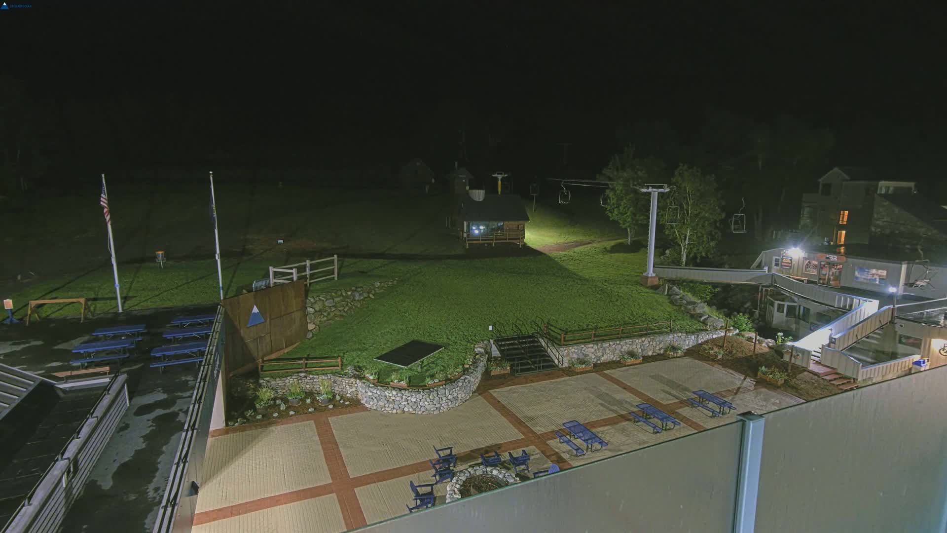 An outdoor nighttime scene shows a paved area with picnic tables and a fire pit, surrounded by landscaping and a ski lift, under dark conditions.