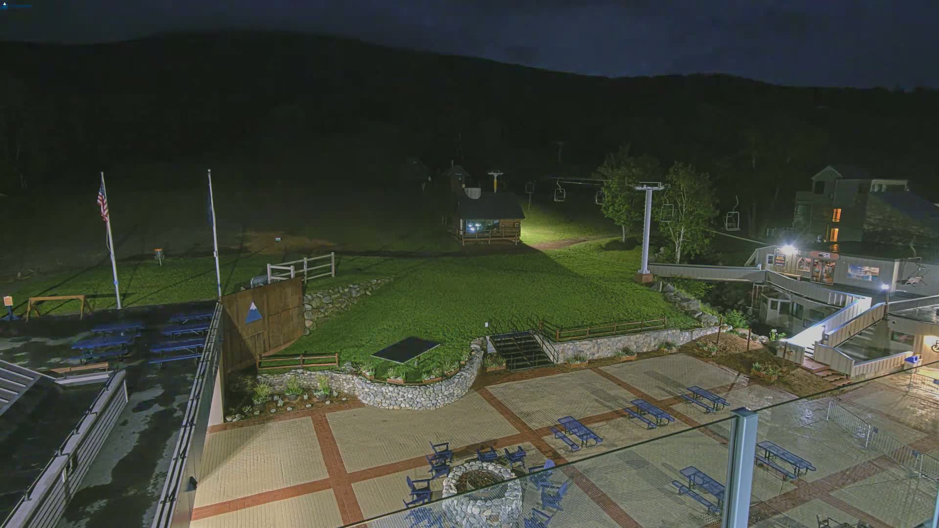 A nighttime view of an outdoor patio area with picnic tables and a fire pit, adjacent to a ski lift and a building, under dark skies.