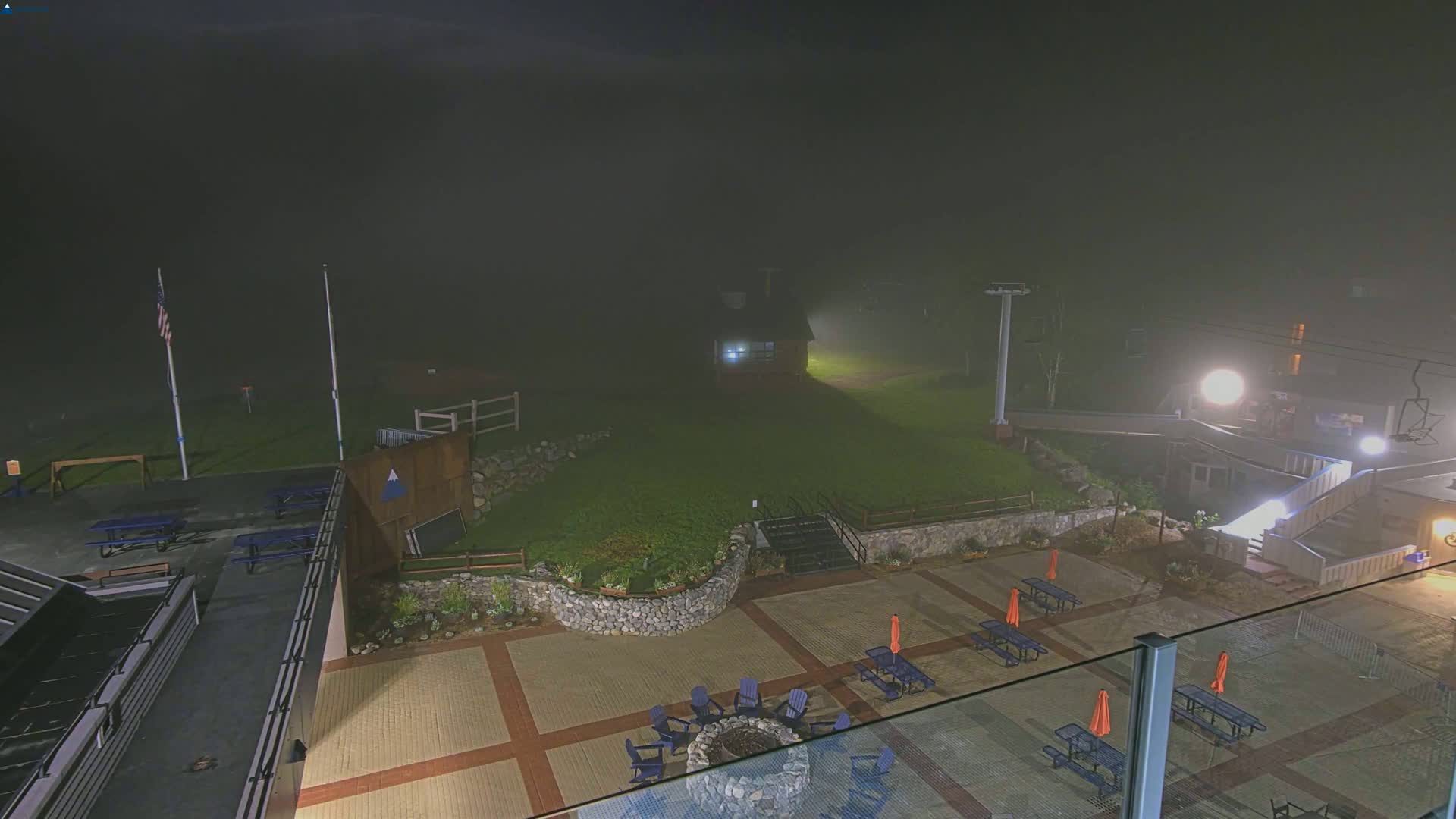 A fog-shrouded outdoor patio area at night, featuring picnic tables, chairs around a fire pit, and a ski lift in the background.