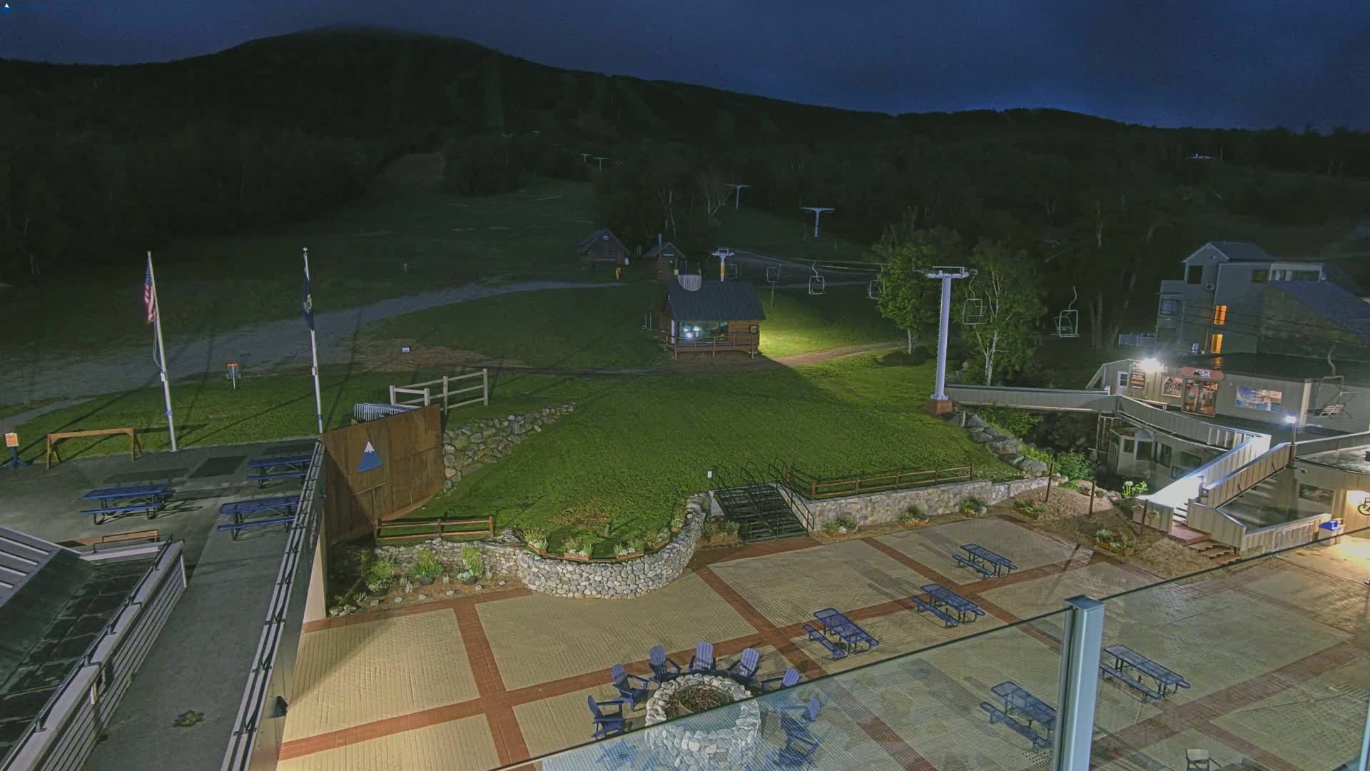 An outdoor patio area with picnic tables and a fire pit is illuminated at night, overlooking a grassy ski slope with chairlifts and a small building under a dark sky.